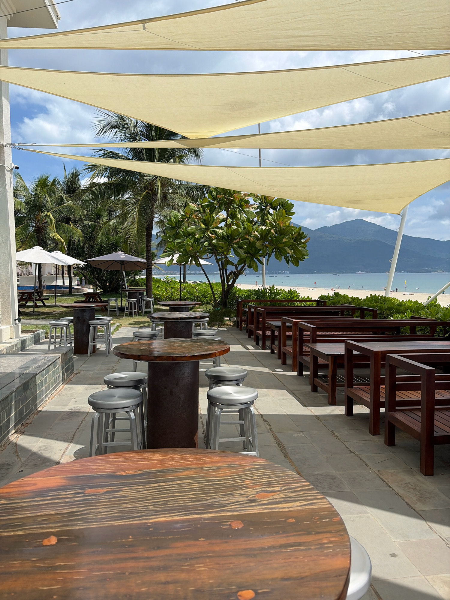 The image depicts an outdoor seating area of a restaurant or cafe situated in Da Nang, Vietnam, with a clear view of a beach and a mountain range. In the foreground, two large, round wooden tables with textured surfaces are visible, surrounded by several metallic bar stools. Behind these, a row of wooden benches and tables forms another seating arrangement. Beyond the seating area, palm trees and lush green foliage create a tropical ambiance. In the middle ground, a beach with people lounging and walking can be seen, with the ocean stretching out to the horizon. The background features a prominent mountain range under a partly cloudy sky. Several white and grey umbrellas are scattered across the scene, providing shade. The architectural element on the left suggests a building adjacent to the seating area, possibly part of the establishment. The overall scene conveys a relaxed, beachside atmosphere.