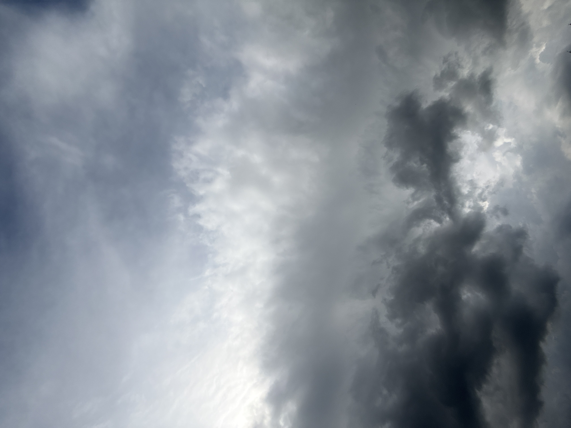 The image displays an overcast sky filled with multiple layers of clouds in the city of Unknown, Unknown. The upper portion of the frame features lighter, wispy cirrus or high altocumulus clouds against a pale blue-gray background, with a bright white light source, likely the sun, diffusing through in the upper-left quadrant. Descending vertically, the cloud cover transitions into a thick, uniform band of medium gray clouds (likely altostratus or stratocumulus). The lower half of the image is dominated by dense, heavy, dark gray to almost black cloud formations, indicative of cumulonimbus or nimbostratus clouds. These lower clouds exhibit significant texture and varied density, with some areas appearing turbulent and lumpy. Patches of lighter gray and white light are visible through gaps in the darker clouds, particularly in the lower-middle and lower-right sections. No terrestrial features, human subjects, or man-made objects are present.