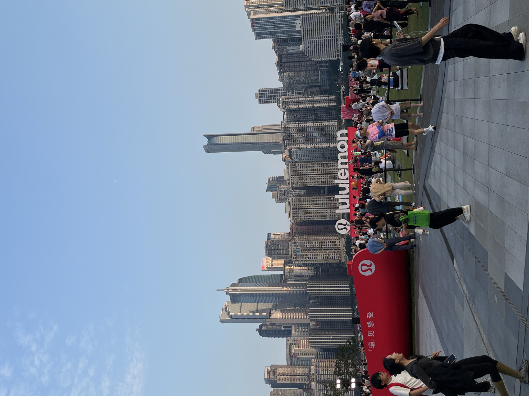 The image depicts a large gathering of people in an outdoor urban setting, with a prominent Lululemon logo and a red, curved structure in the foreground. In the background, a dense cityscape with numerous high-rise buildings is visible under a clear blue sky. The red structure appears to be an inflatable or fabric installation, possibly part of an event or promotion, with Chinese characters and the Lululemon logo displayed on it. People are casually dressed and spread out across a paved area and a grassy section in the midground. Some are walking, while others are sitting or standing, seemingly observing or participating in an activity. The overall atmosphere suggests a public event, possibly a fitness or community gathering, taking place in a modern city. The city in the background is characteristic of Jiangbeicheng Street (江北城街道), Chongqing, China, known for its modern architecture and dense urban development along the riverfront.