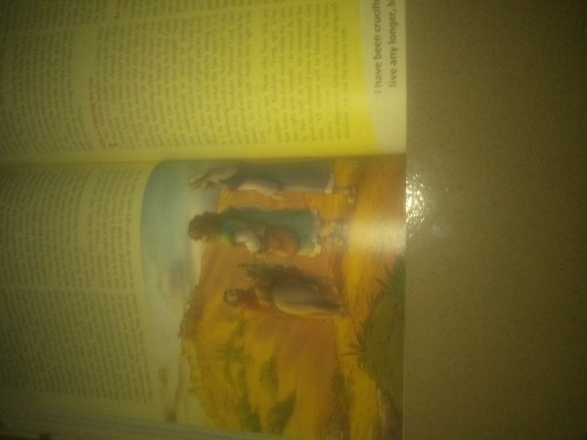 The image shows an open book with text on the left page and an illustration on the right page. The text on the right page is partially obscured but includes the phrase "I have been crucified, live any longer, b". The illustration depicts three figures in a landscape with a mountain. One figure, in the foreground, is seen from behind, appearing to carry something on their back. Two other figures are standing or seated above and to the left of this figure. The image is taken from a top-down perspective, and the lighting is uneven, suggesting an indoor setting with a light source from above and to the right. There is no visible information within the image to suggest a specific location in Jauro Yinu, Nigeria.