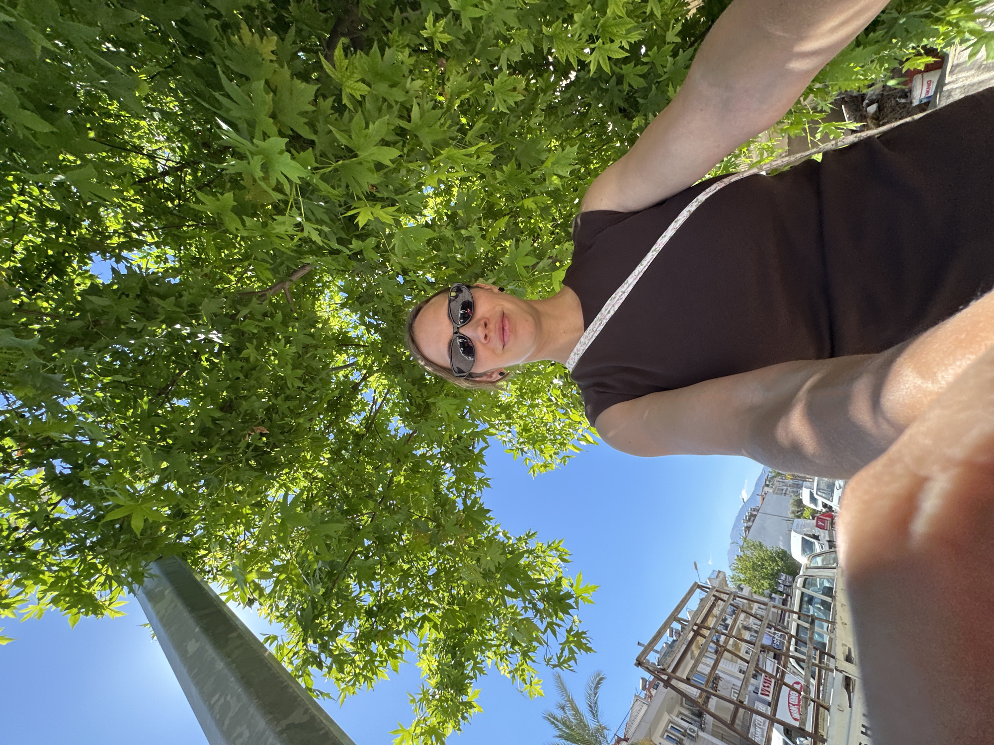 The image shows a woman taking a selfie while standing under a tree in Fethiye, Turkey. The camera is angled upwards, capturing her face, part of her brown dress with a white strap, and her arms. She is wearing sunglasses. The tree, with its dense green leaves, forms a canopy overhead, with patches of blue sky visible through the foliage. In the background, buildings and vehicles are visible, indicating an urban setting. A tall, metallic pole stands to the left of the frame. The image seems to be taken during the day, under bright sunlight.