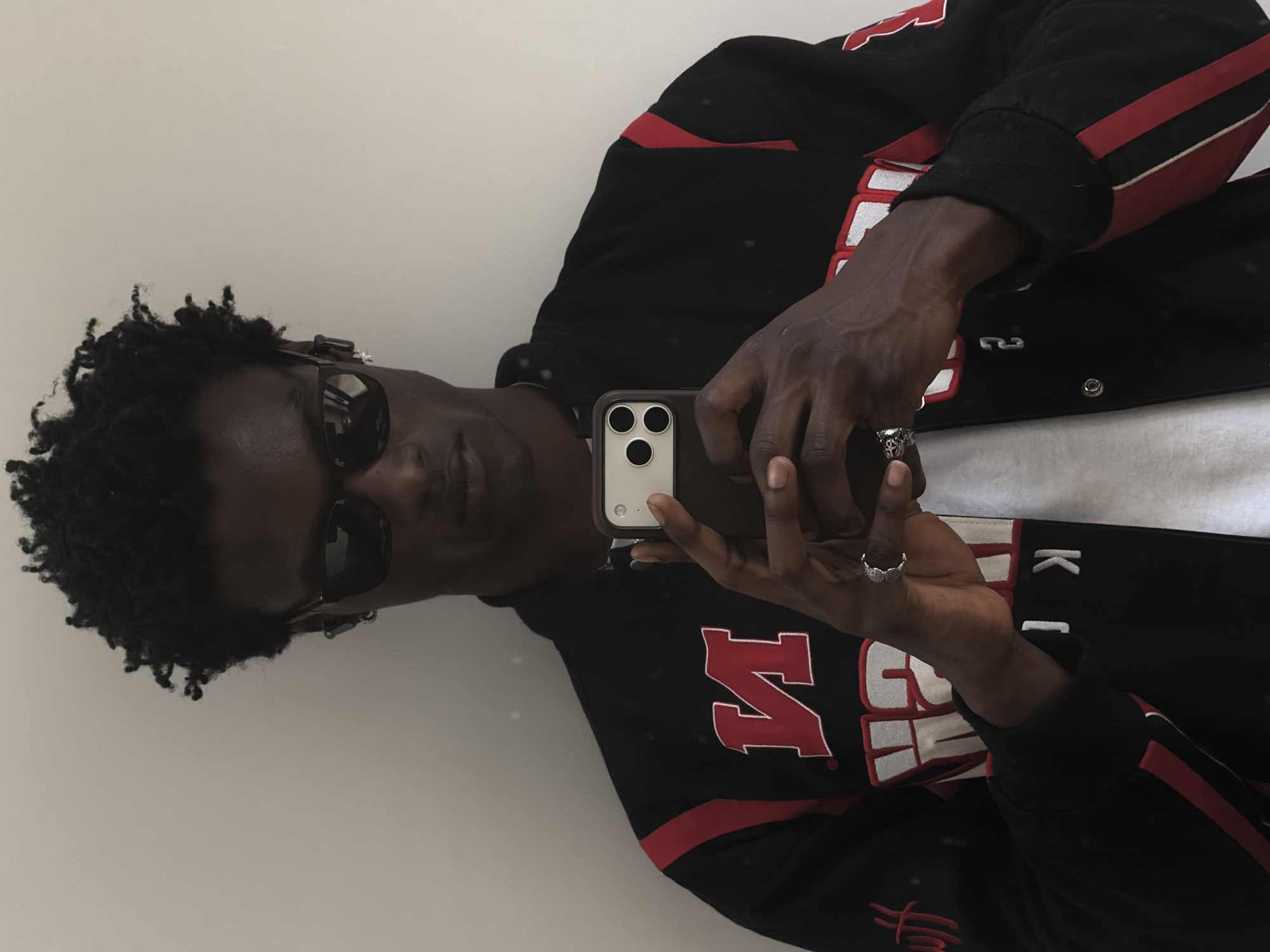 A dark-skinned person wearing sunglasses takes a selfie with a smartphone. They are wearing a black jacket with red and white detailing. They are also wearing rings on their fingers. The background is plain white.