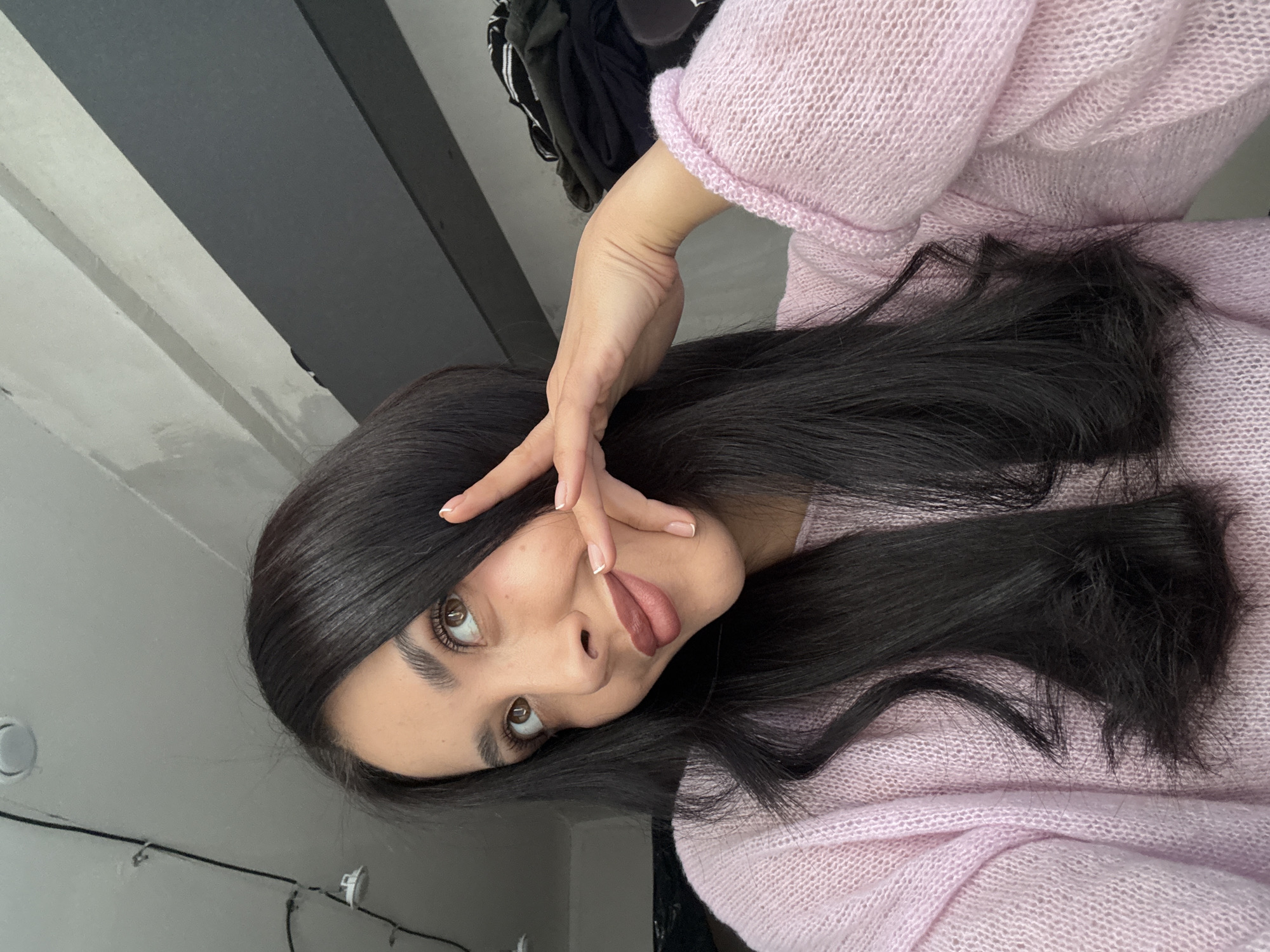 A young woman with long, straight, dark hair is posing for a selfie. She is wearing a light pink, knitted sweater. She has fair skin, brown eyes, and defined eyebrows. Her lips are coated in a matte, mauve-colored lipstick. She is holding one hand up near her face, with her fingers spread apart. The background is a mix of gray wall and ceiling. The location in the city of Votkinsk, Russia cannot be determined from the image.