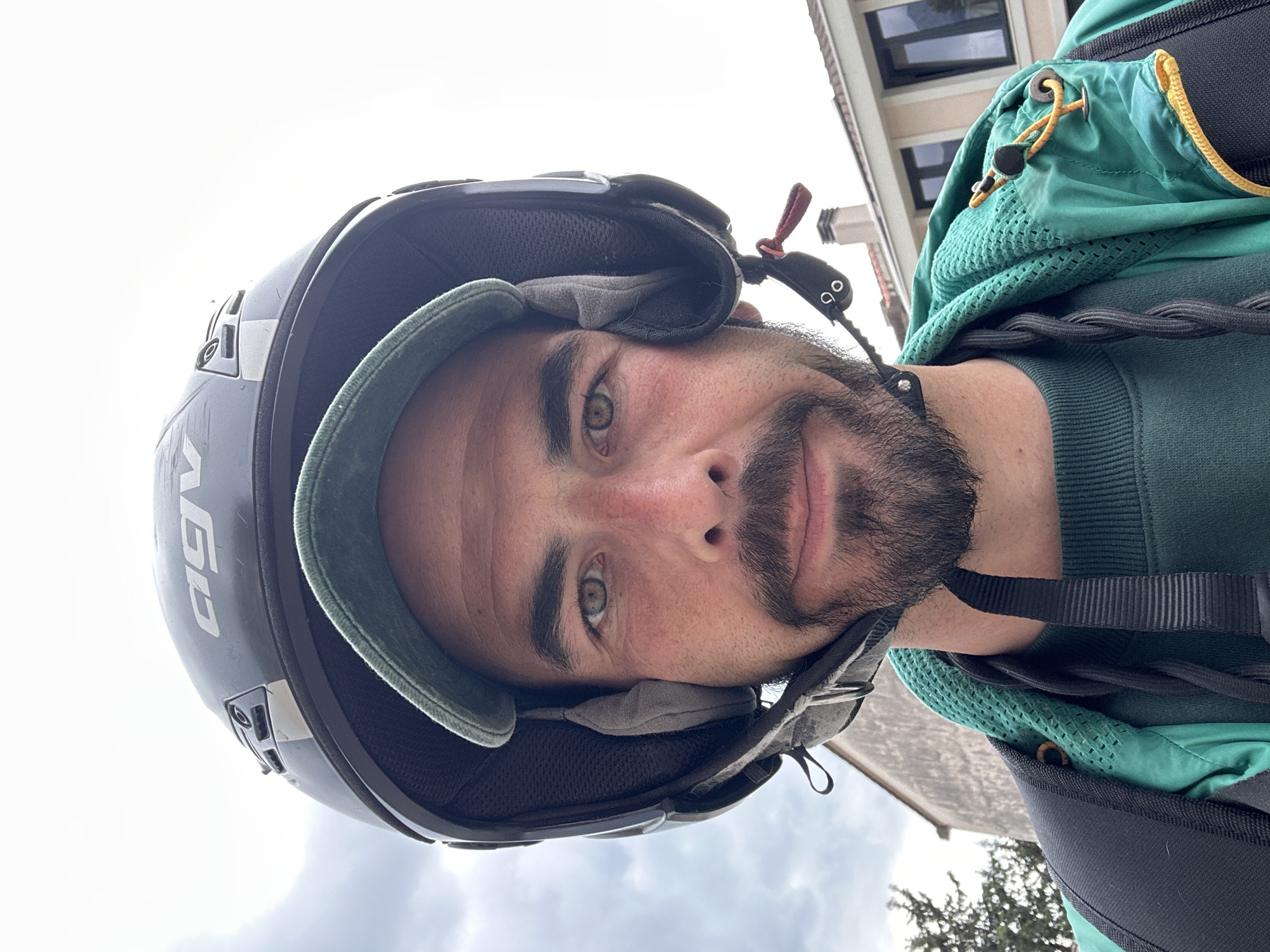 A man with a beard and green eyes is wearing a black helmet. He is also wearing a green jacket and sweater. The helmet has "AGV" written on the side. He appears to be outdoors with a building visible in the background. The location cannot be confirmed as the city of Budva, Montenegro.
