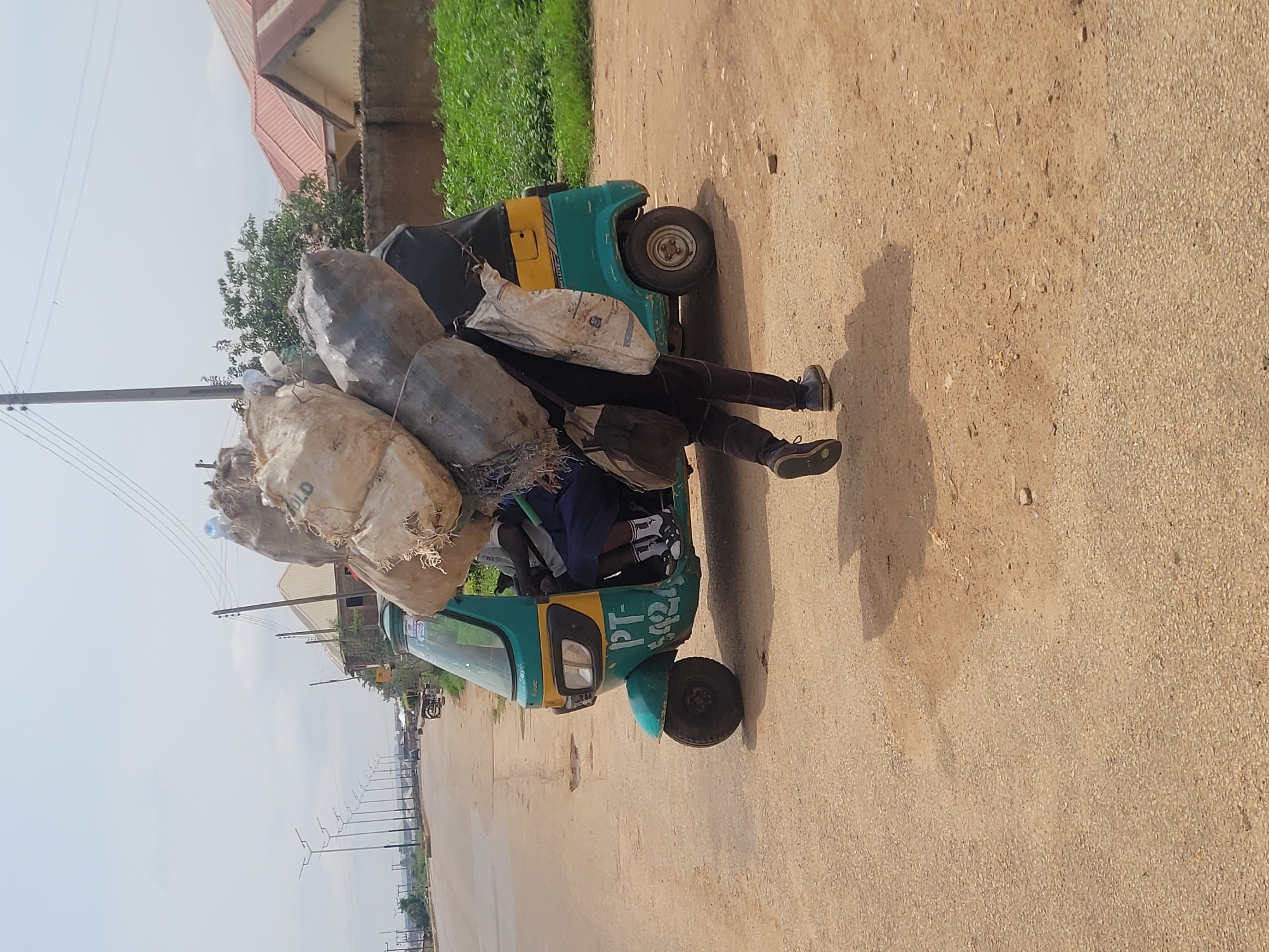 A person is walking alongside a teal and yellow auto rickshaw, carrying a large load of bundled sacks and plastic bags on their back. The rickshaw has the markings "PT-5427" on its side. The person is wearing dark pants and dark shoes, and appears to be a male. The auto rickshaw is stationary on a dusty road, with the driver visible inside. In the background, a row of streetlights stretches down a paved road, with buildings and greenery on the right side. This scene is likely in Du, Nigeria, given the context of the uploaded image. The sky is overcast, suggesting a cloudy day.
