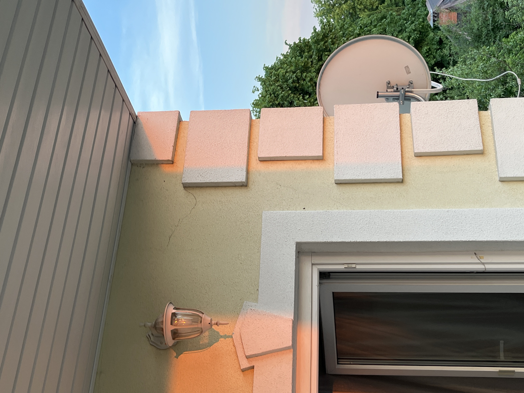 The image shows a section of a building with a light yellow exterior wall under a blue sky with some trees visible at the top. The wall has a decorative series of square protrusions along the top, which are a lighter color than the wall. A white satellite dish is mounted to the wall near the top. A white-framed window is visible, partially open. An outdoor light fixture is mounted on the wall. A white, vertically-lined siding is visible to the left of the wall. The location is Unknown, Unknown.