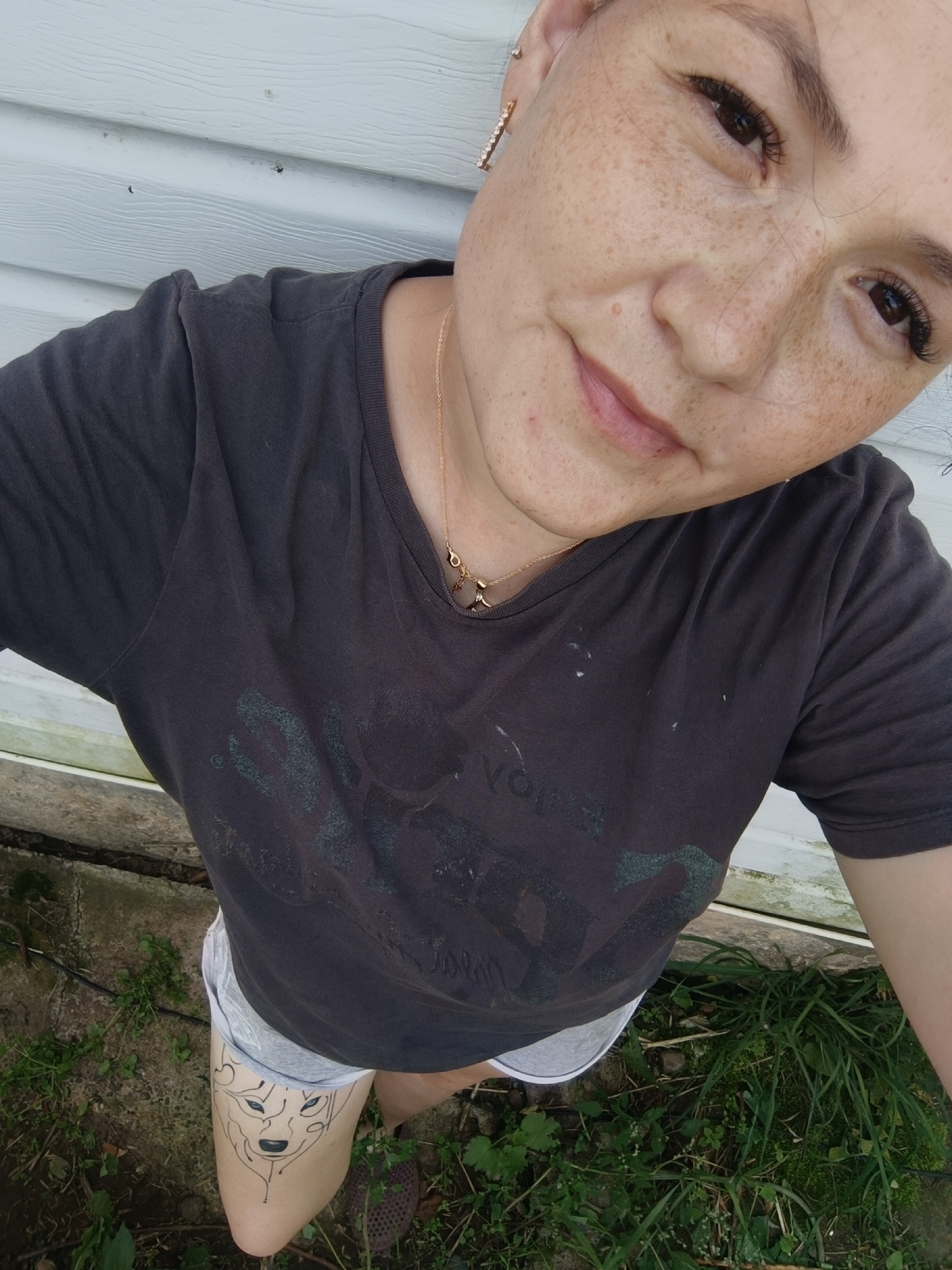 The image shows a woman with freckles wearing a dark t-shirt and light-colored shorts. She has a necklace with charms and earrings. On her left thigh, there is a tattoo of a wolf's face. She is standing outdoors in what appears to be a backyard, with green grass and a white siding wall in the background. The location is not specified as being in the city of Karsha, Russia.