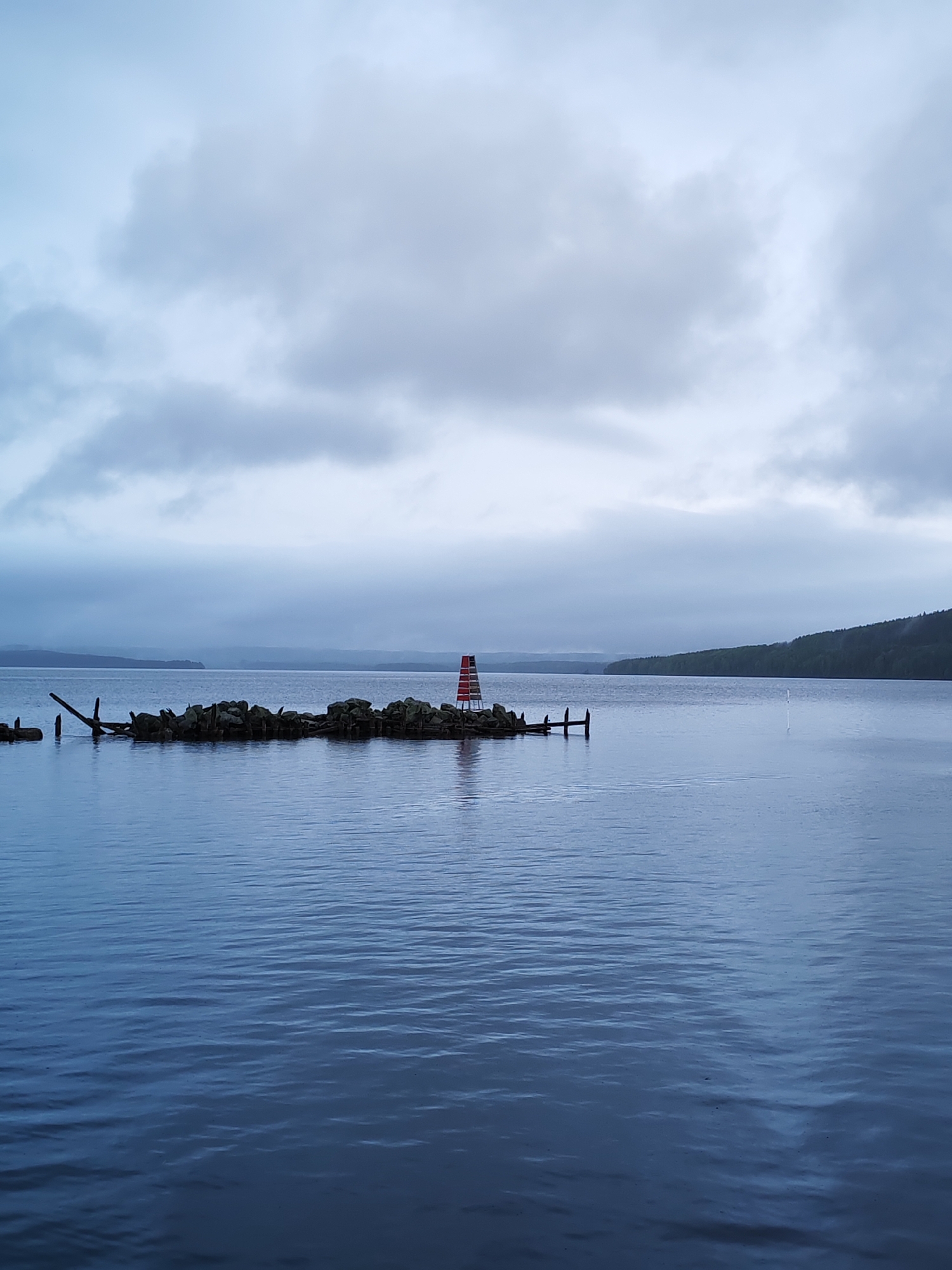 The image presents a serene and expansive landscape dominated by a large body of water under a heavily overcast sky. In the mid-ground, a small, rocky island or breakwater stretches across the water, constructed from dark rocks and weathered wooden debris. A prominent feature on this island is a striking red and white striped structure, resembling a navigational beacon or a small lighthouse, which stands vertically with distinct red and white bands.
Distant forested shores or hills frame the far edges of the water on both the left and right, receding into a hazy horizon and suggesting a vast lake or wide river. The sky is completely covered by thick, grey clouds, indicating an overcast day and casting a soft, diffused light over the entire scene. The water surface is a deep blue, textured by gentle ripples that reflect the subdued light of the sky.
No people or explicit activities are visible, contributing to a tranquil and solitary atmosphere. The overall impression is one of calm and natural beauty on a cloudy day. Given the context, this scene likely depicts Lake Onega near Medvezhyegorsk, Russia. There is no visible text in the image.