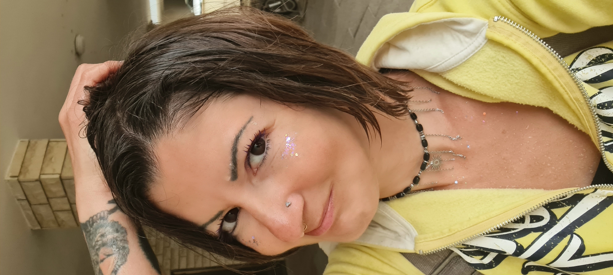 A person with short, dark, wet hair is seen from the chest up, tilted to the right, with their right hand near their head. They are wearing a yellow jacket. There is pink glitter on their face and chest. They are also wearing a black and silver necklace. They have a nose piercing in each nostril. There is a tattoo visible on their left arm. The background is indistinct. The image does not show any identifying information to suggest a specific location like Nahariyya, Israel.