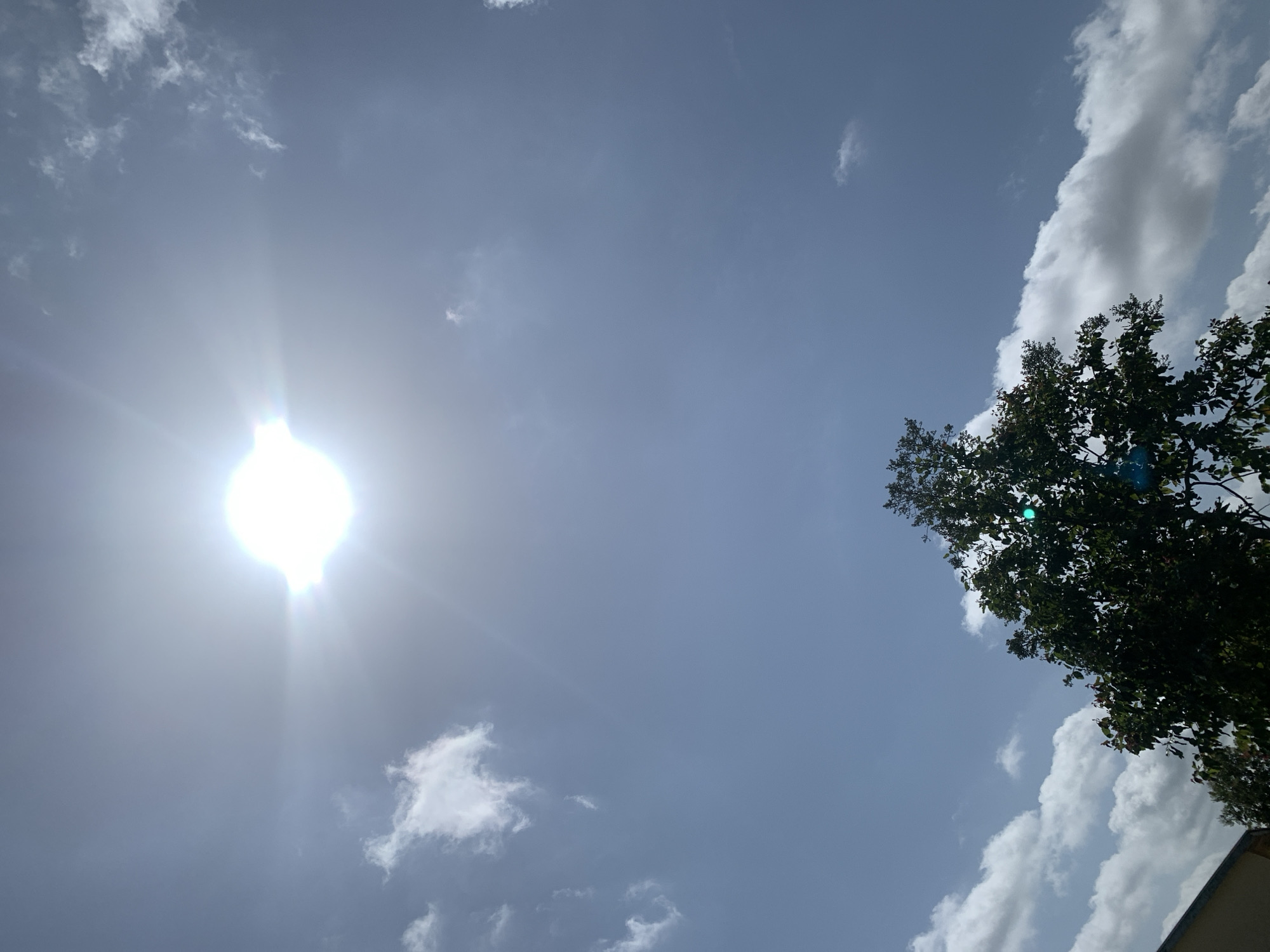 The image is an outdoor, low-angle shot of a bright sun in a blue sky with scattered clouds. A portion of a green leafy tree is visible in the lower part of the frame, along with the roofline of a building on the far left. The sun appears in the upper center of the image and exhibits a lens flare effect, casting rays across the sky. The scene depicts a clear, sunny day. The location is identified as Kuru Karama, Nigeria. There are no people, animals, or specific actions depicted in the image.