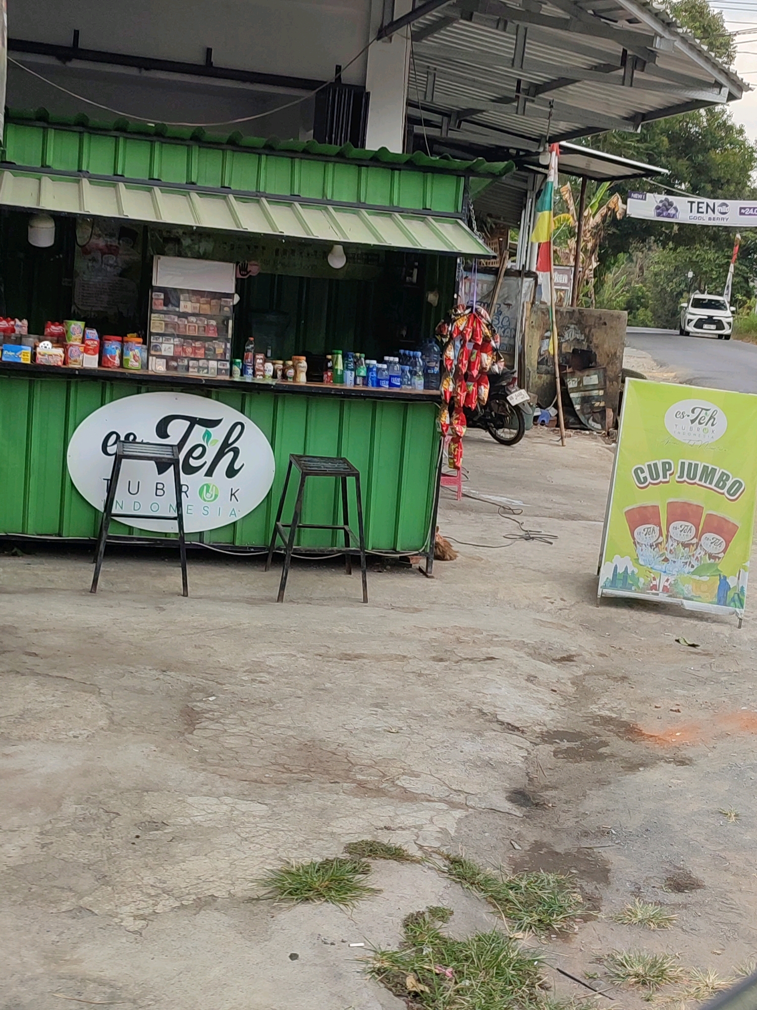 This is a medium-shot, eye-level, outdoor, daytime photograph taken in Sambak, Indonesia. The foreground is a concrete surface with patches of grass and some debris. In the midground, a green food stall is set up, with a counter displaying various packaged snacks and beverages. Behind the counter, a refrigerator unit is visible, filled with drinks. Two dark metal stools are placed in front of the stall, presumably for customers.
A large, oval-shaped sign with the text "es Teh Tubruk Indonesia" is mounted on the green stall. To the right of the stall, a brightly colored, green A-frame sign advertises "CUP JUMBO" with images of iced tea cups. In the background, a white car is driving on a road, and lush green trees and vegetation line the road. A flag with yellow, red, and green stripes is planted near the stall. A motorcycle is parked behind the stall, and various bags of snacks are hanging from a rack. A sign with "TEN miles" and images of blueberries is visible in the upper right portion of the image. The overall scene depicts a roadside food stall in a semi-rural or suburban setting in Indonesia.