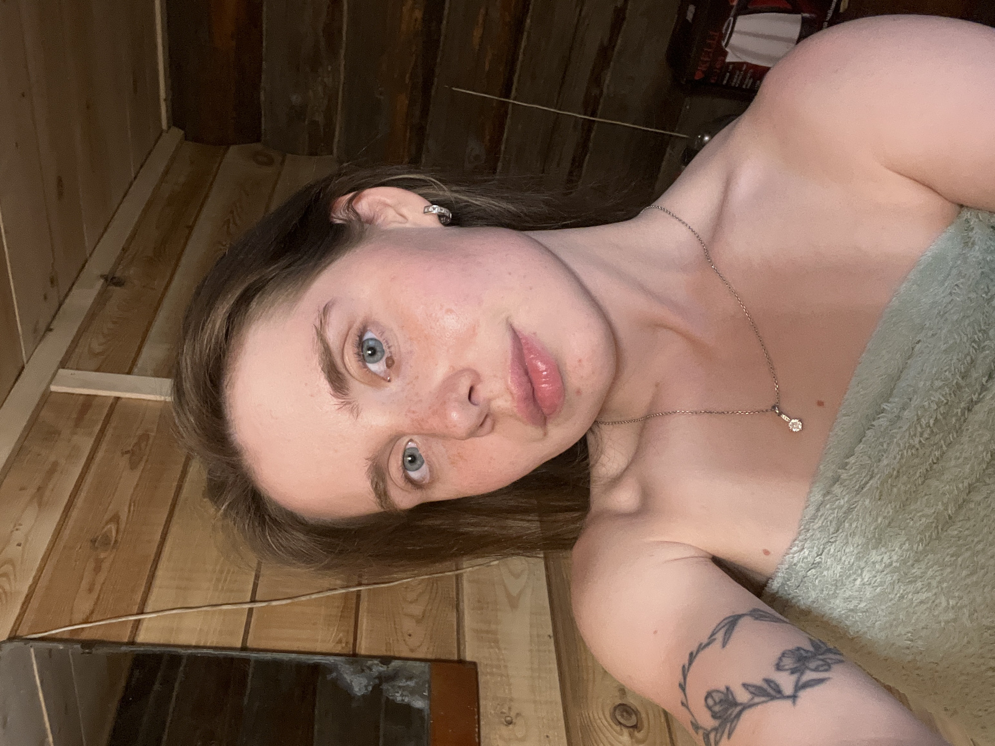 A fair-skinned woman with light brown hair and blue eyes is photographed at close range. She is wrapped in a light green towel and wearing a delicate silver necklace with a small pendant. A floral tattoo is visible on her left upper arm. The background consists of wooden paneling and a dark metallic object in the lower left corner. The location is a sauna, likely in Chaikovsky, Russia, as requested.
