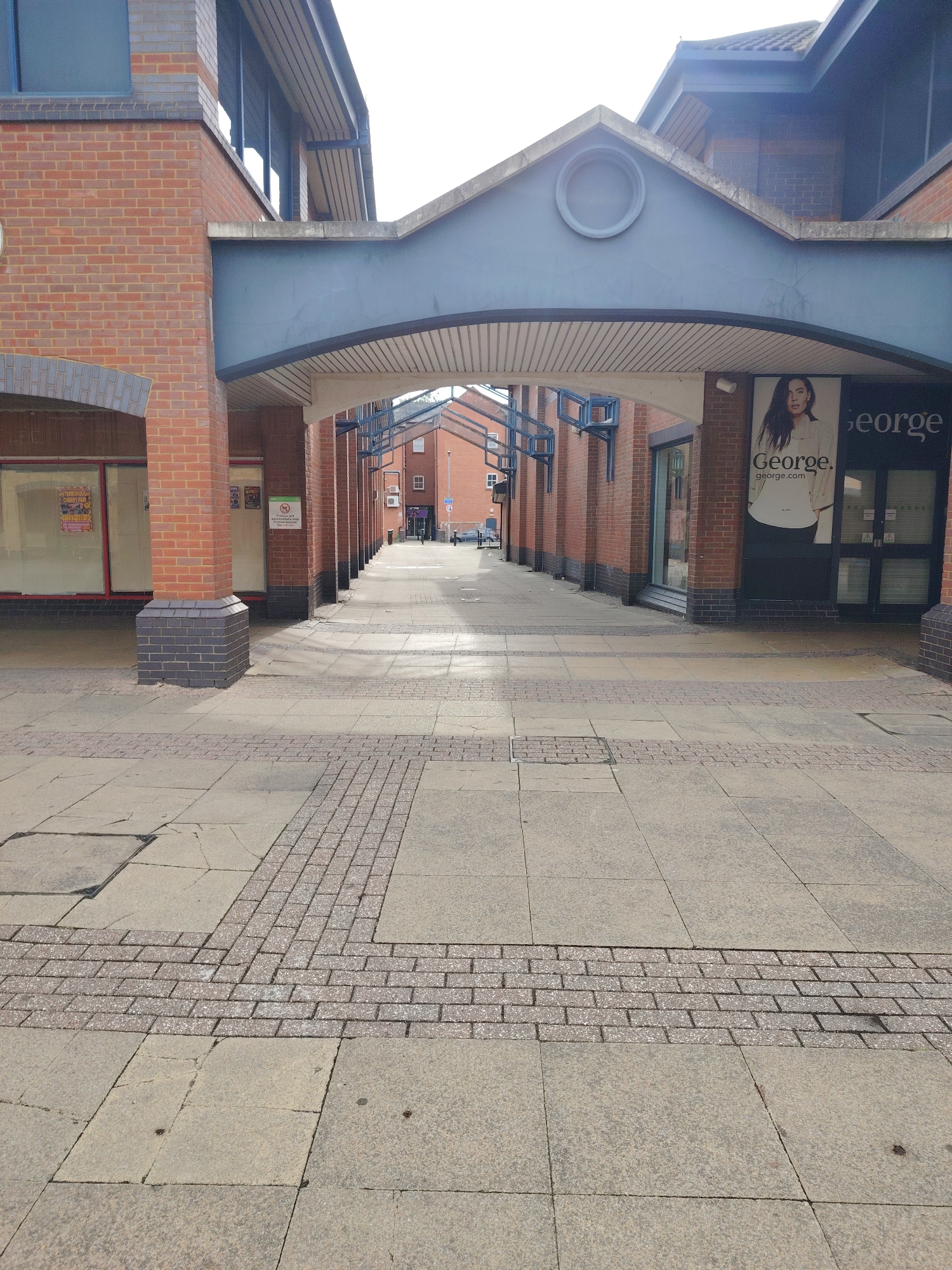This image depicts an outdoor pedestrian walkway or precinct in the city of Peterborough, United Kingdom, seen on a bright, sunny day with clear shadows. The perspective is looking down the length of the passage.
The foreground features a paved ground composed of large, light grey concrete slabs interspersed with decorative sections of smaller, reddish-grey rectangular pavers. These decorative sections form a cross-like pattern near the viewer and a linear strip further into the scene. Minor cracks and wear are visible on the concrete slabs.
The walkway is bordered on both sides by multi-story brick buildings featuring prominent red brickwork with darker grey/blue brick bases. These buildings are supported by evenly spaced brick pillars along the pedestrian route. An overhead structure, painted in a muted blue-grey, forms a distinctive arched canopy spanning the passage. The underside of this arch is lined with light-colored slats. Above the arch on the right side, a circular window or decorative element is integrated into the structure.
On the right side of the passage, a large retail shopfront displays a prominent "George." logo and "george.com" alongside a large image of a female model. Other shopfronts are visible further down the passage. On the left side, one visible shopfront appears vacant, displaying various posters internally. A white sign on a pillar on the left indicates that "Smoking or vaping is not permitted in this area."
In the distance, the passage extends between more brick buildings, with a light-colored vehicle partially visible at the far end. No people are present in the immediate or distant view of the scene. The architectural style, shop branding, and overall environment are consistent with a commercial pedestrian area in a UK town or city.
