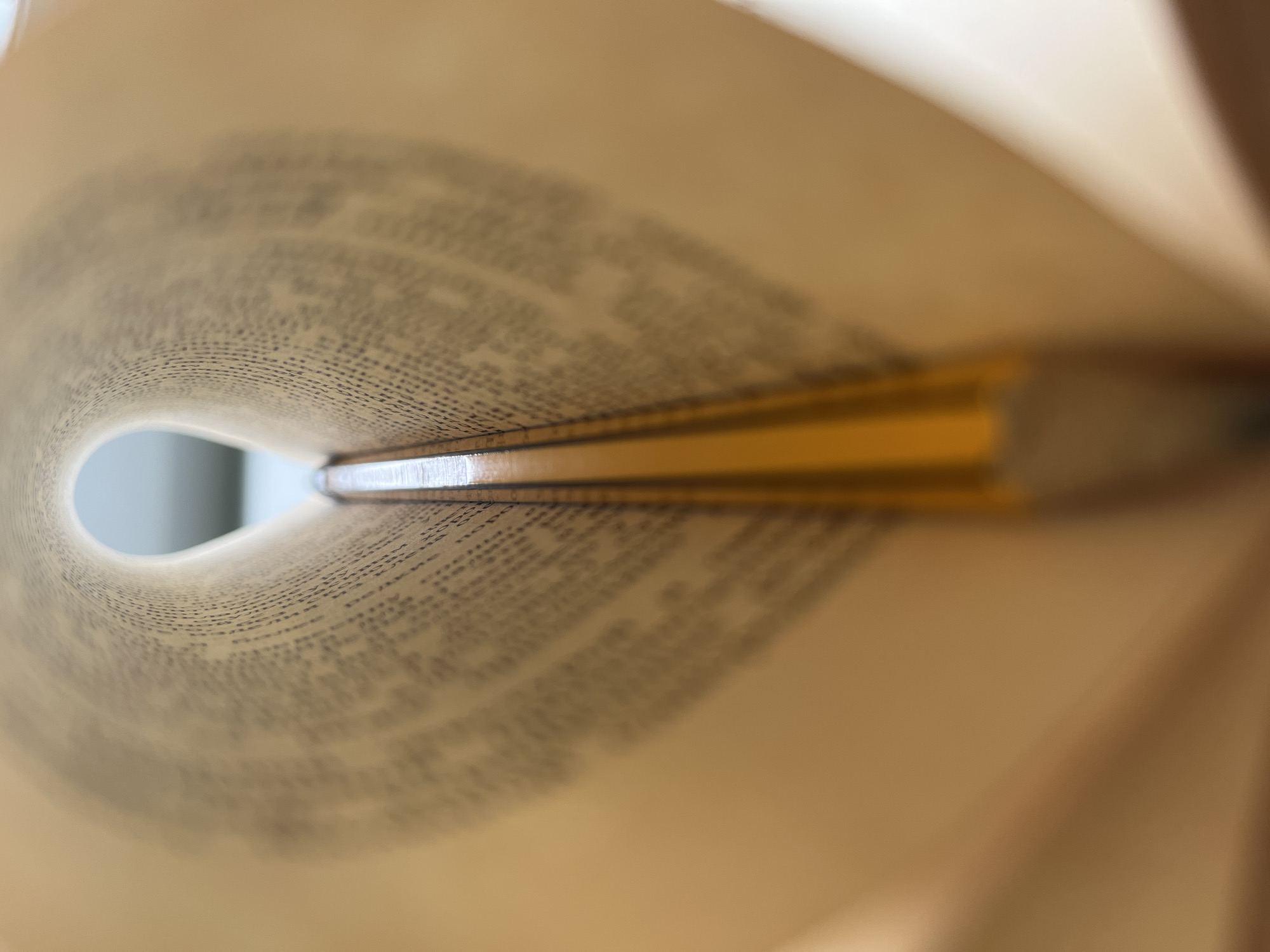 The image shows a close-up, perspective view into the center of a rolled-up book. The pages form a tunnel shape, with printed text visible on the inside surfaces. The book's spine is visible in the middle ground. A blurred white area is seen through the opening at the far end of the tunnel. The book is pale yellow. The location and city information are not discernible from the image.