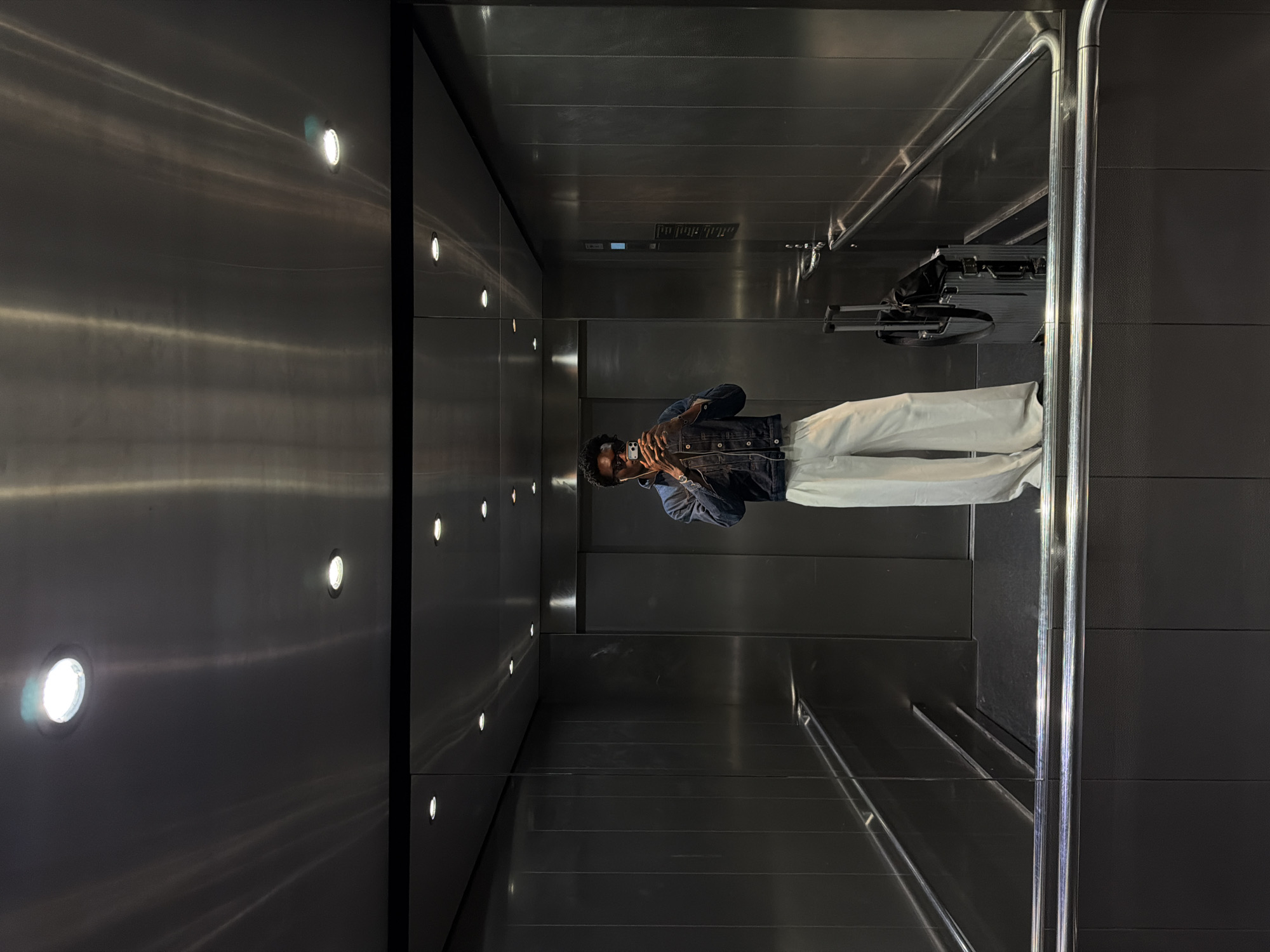 The image depicts the interior of an elevator in Crawley, United Kingdom. The elevator walls are constructed of dark gray, metallic paneling, punctuated by rows of small, circular lights. A person, appearing to be male, stands inside the elevator, facing the mirrored wall. They wear white pants and a dark denim jacket and are holding a mobile phone up as if taking a photo in the mirror. Behind them, luggage with a pull handle sits on the elevator floor. Grab bars are positioned near the edge of the mirror. The elevator floor is also paneled in the same dark gray metallic material.