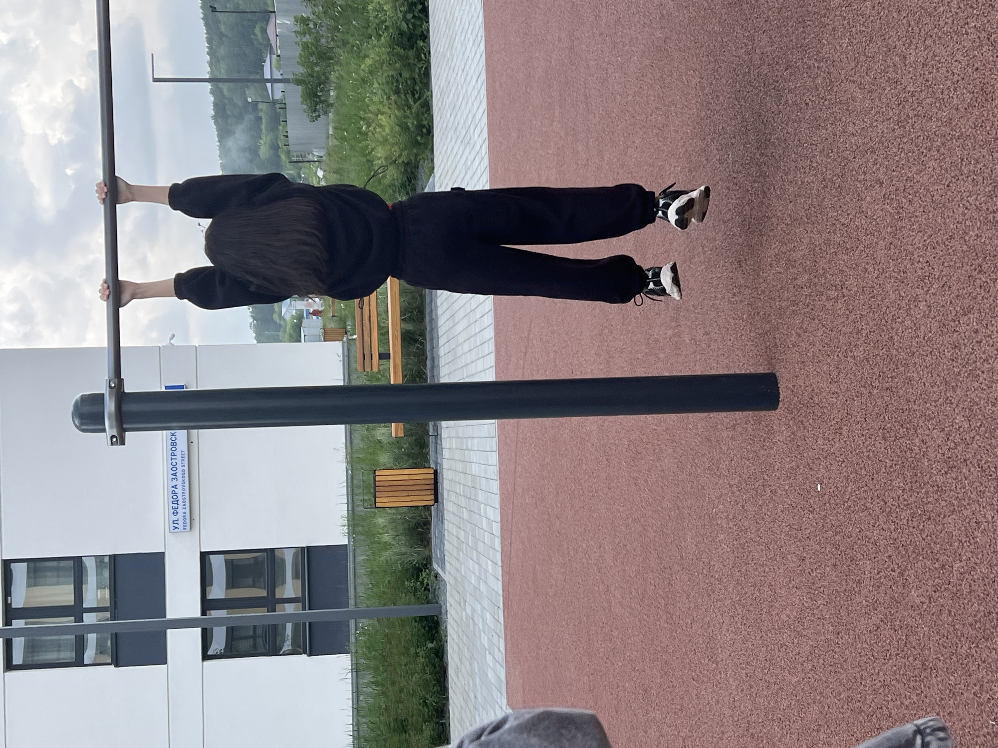The image depicts an outdoor scene on Fedora Zaostrovskogo Street in Yekaterinburg, Russia, featuring a person hanging from a horizontal bar. The primary subject, seen from the back, is a young individual with long, straight, dark hair, wearing a black long-sleeved top and black sweatpants, suspended in the air by their hands gripping a dark gray horizontal bar. Black and white athletic shoes are visible on their feet.
The horizontal bar is supported by a dark gray vertical post on the right; the left support is out of frame. Beneath the bar and extending into the foreground is a reddish-brown, granular, soft-surface ground cover, likely rubberized. To the left, a partial view of a gray garment indicates the presence of another individual outside the main frame.
In the midground, behind the person and the bar, a paved area with light and dark gray rectangular tiles is visible. To the right of this paved section, a wooden slatted bench is present, and further back, a wooden trash receptacle with a black frame stands near the edge of the paved area. Green vegetation, consisting of shrubs and tall grasses, borders the paved surface.
The background features a multi-story building with a white facade and dark gray window frames, to which a blue street sign is affixed, reading "УЛ. ФЕДОРА ЗАОСТРОВСК" (FEDORA ZAOSTROVSKOGO STREET). Beyond the immediate activity, a landscape of distant trees and some structures under an overcast sky is visible, with faint smoke or haze rising from behind the trees on the left.