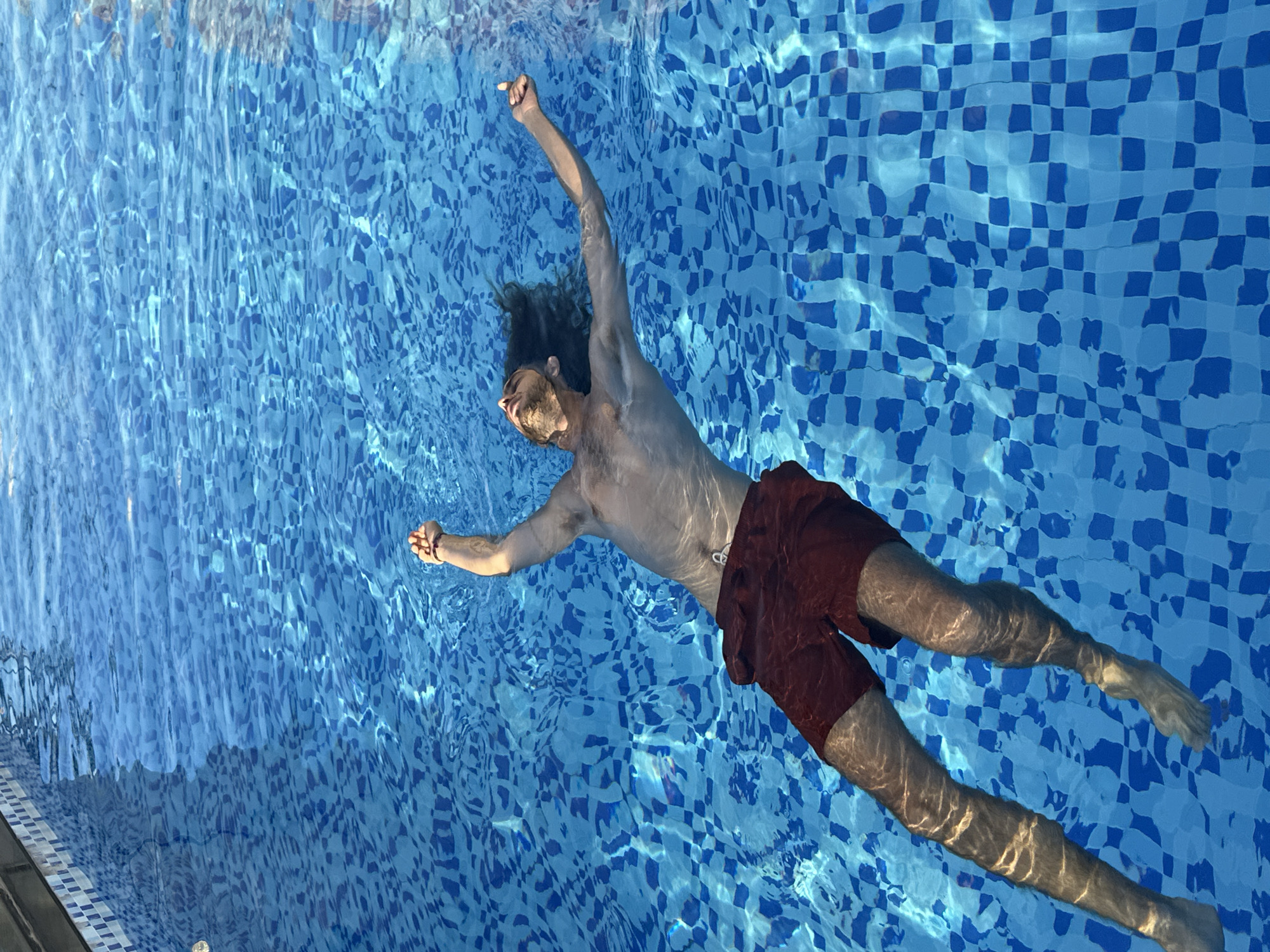 A man with long, dark, wavy hair floats on his back in a swimming pool. He is wearing dark red swim trunks. His arms are extended above his head, and his face is turned upwards, eyes closed, with a relaxed expression. The water is clear blue, with sunlight reflecting off the surface, creating a shimmering effect on the tiled bottom of the pool. The pool appears to be an outdoor setting, possibly a hotel or residential complex, given the tiled edge visible in the upper left corner. The location is Đà Nẵng, Vietnam.