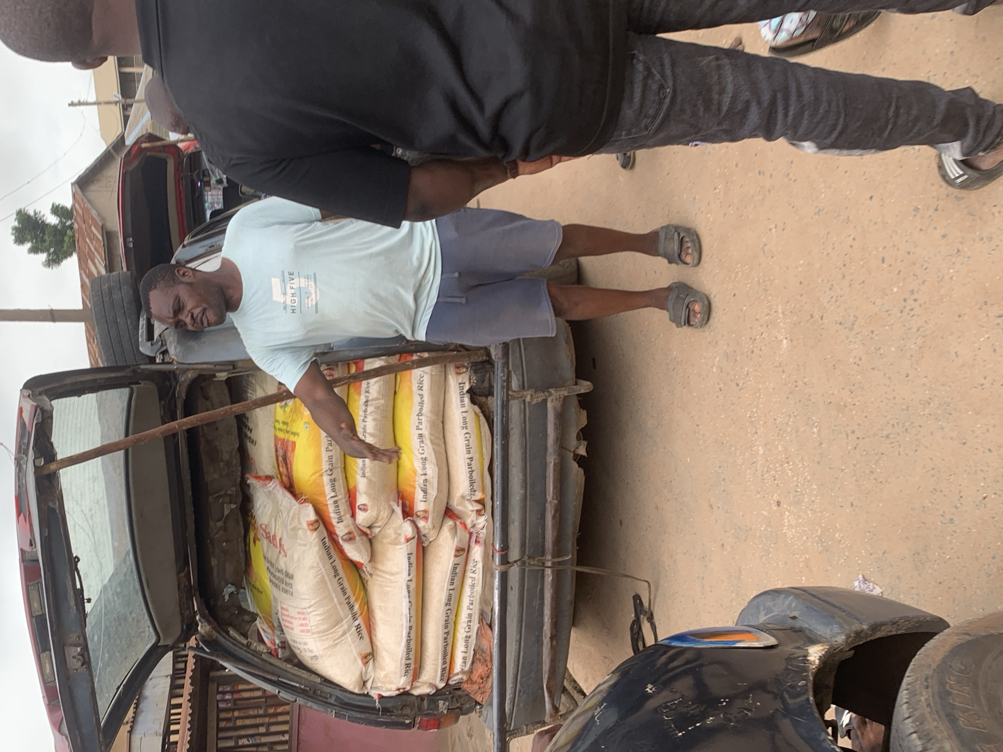 The image captures a scene in Shagamu, Nigeria, focusing on a man standing next to an open vehicle loaded with sacks of rice. The vehicle, possibly an SUV or van with its rear hatch open, is positioned on a dirt road. The sacks are predominantly yellow and white, with visible text indicating "Indian Long Grain Parboiled Rice." The man in the foreground is wearing a light blue t-shirt with "HIGH FIVE" printed on it and grey shorts, with grey sandals on his feet. He is gesturing towards the back of the vehicle with his right hand. Another individual, wearing a black t-shirt and dark denim jeans, stands to the right, his back mostly to the camera. The background shows a row of buildings with corrugated iron roofs, suggesting a residential or commercial area in Shagamu. The overall environment appears somewhat dusty and utilitarian.