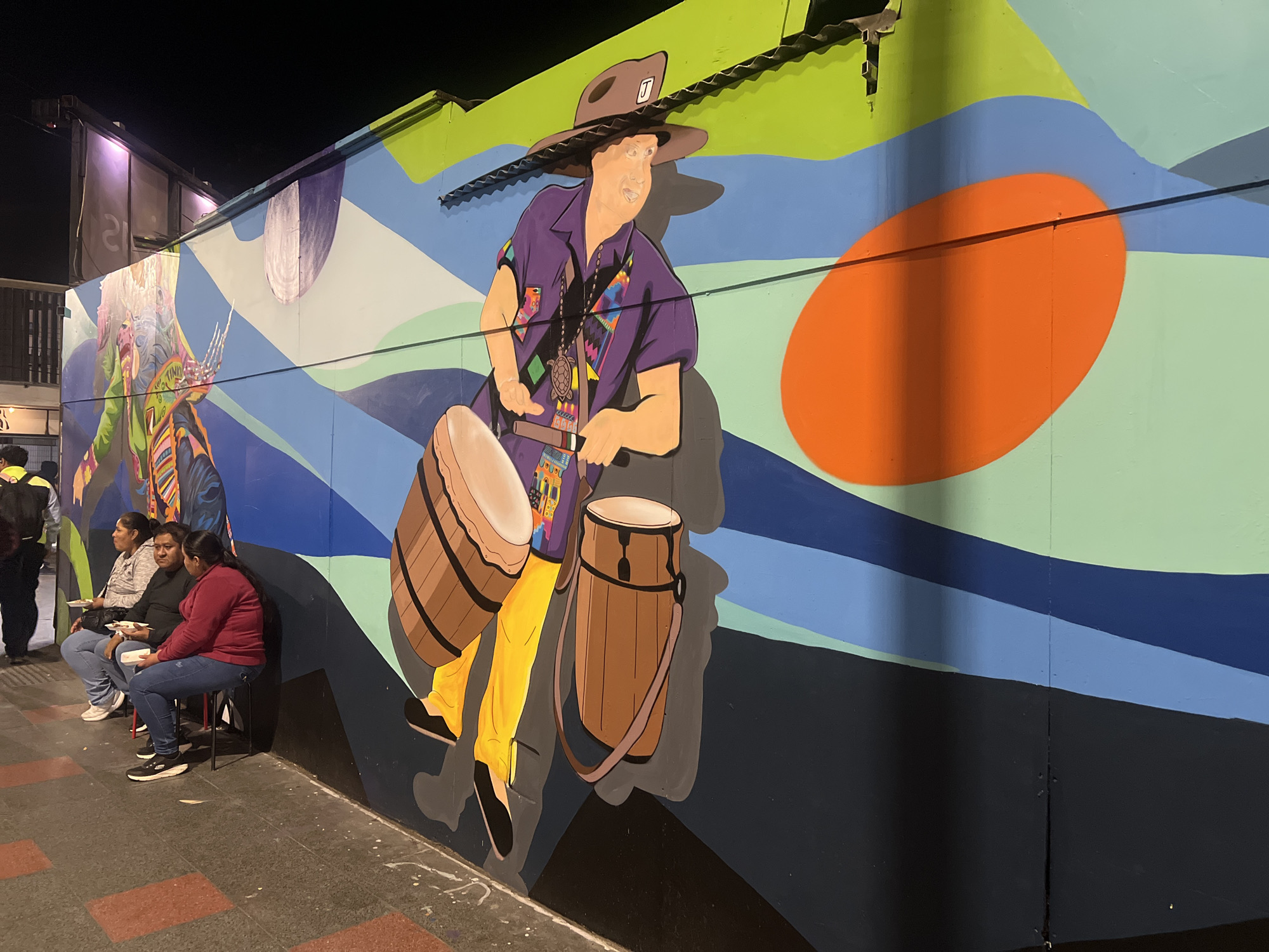The image shows a brightly colored mural on the side of a building in Arica, Chile. The mural depicts a person in a purple shirt and brown hat playing drums. In front of the mural, three people are seated on chairs, eating from white plates. The ground in front of the mural is paved with red and gray square tiles. A street artist is walking away from the viewer in the background. The mural is abstract and colorful, with shades of blue, green, and orange. The people are casually dressed and appear to be enjoying their meal. The overall scene is one of urban life in a South American city.