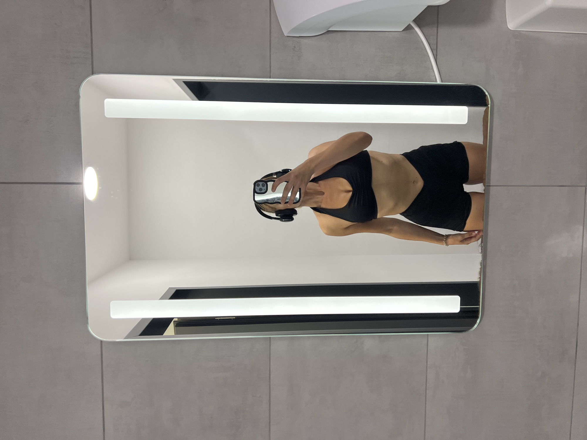 A woman wearing a black sports bra and black shorts stands in front of a mirror, taking a selfie with her phone. She also has black headphones on. The mirror is illuminated by two vertical lights on either side and a circular light above. The background is a gray tiled wall. The scene appears to be in a restroom or locker room, possibly at a gym. There is a white hand dryer mounted on the wall to the right of the mirror. The image does not provide any specific information about the location in the city of Altötting, Germany.