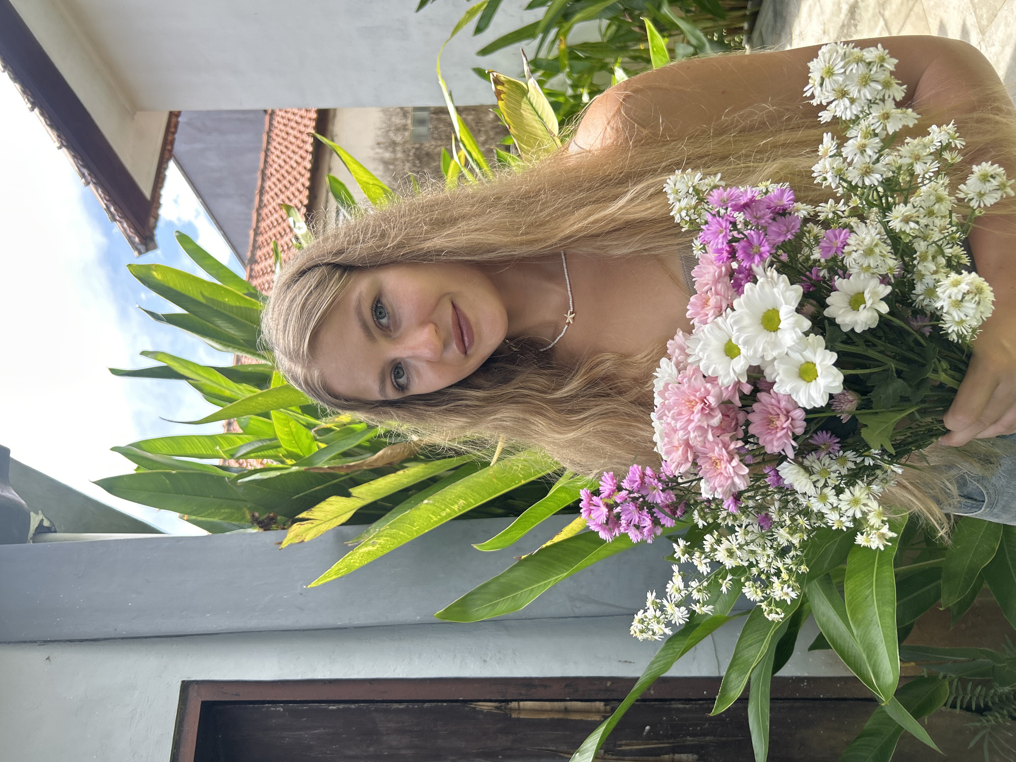 The image is oriented on its side, rotated 90 degrees counter-clockwise. It features a young woman with long, wavy blonde hair, set outdoors in Kuta Selatan, Indonesia. Her head is slightly tilted, and she looks directly at the viewer with a gentle, relaxed smile. Around her neck, she wears a delicate necklace featuring a small white bead and a gold star charm.
She holds a large, colorful bouquet of fresh flowers, predominantly featuring white, various shades of pink, and purple blossoms, resembling chrysanthemums, daisies, and asters. The vibrant bouquet occupies a significant portion of the lower right side of the frame as seen in the rotated image, held against her upper body. Her hand is visible holding the stems near the bottom of the bouquet.
The background depicts a bright, sunny day with a clear blue sky and a few wispy clouds visible in the upper left. Lush, broad-leafed green plants, some illuminated by sunlight, are prominent behind the woman and the flowers. Architectural elements of a building with a distinctive terracotta-tiled roof are visible in the upper left part of the background. A grey concrete wall or ledge runs horizontally across the mid-ground. The overall atmosphere conveyed is cheerful and natural, suggesting a moment captured during the morning or late afternoon. There is no visible text in the image.