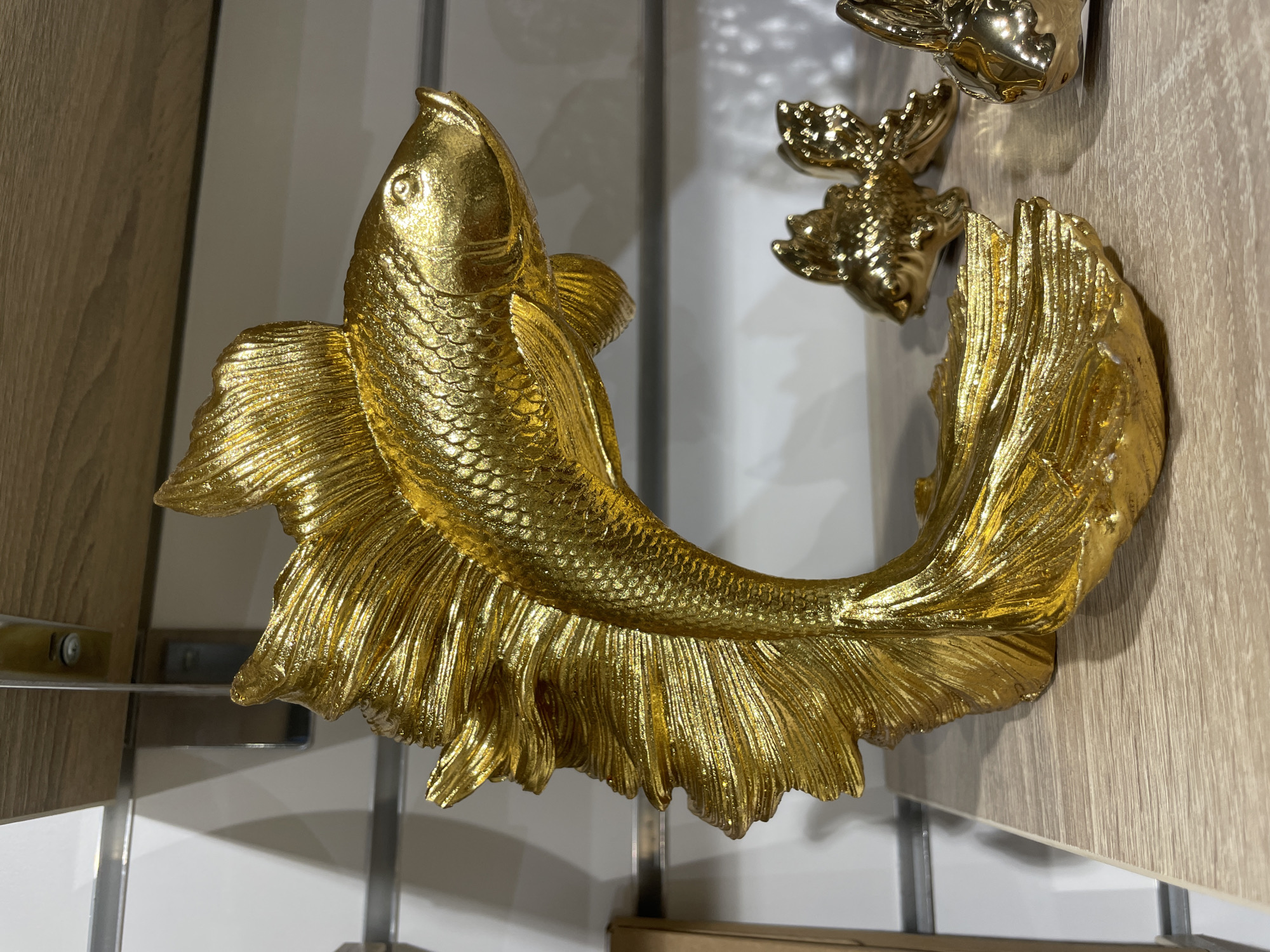 A gilded fish figurine is displayed on a wooden shelf. The figurine is a decorative object, likely made of ceramic or resin with a gold-tone finish, shaped to resemble a betta fish with flowing fins. It is positioned on the left side of the frame, with its body curved and its tail fanned out. In the background, to the right, are two smaller, similarly gilded fish figurines. The shelf is made of light-colored wood with a visible grain and is part of a display unit with metal shelving supports and a white, textured wall behind it. The scene appears to be an interior shot, possibly in a retail setting or home decor display. There are no people or any indication of activity. The image does not provide any information or context related to the city of Perm, Russia.