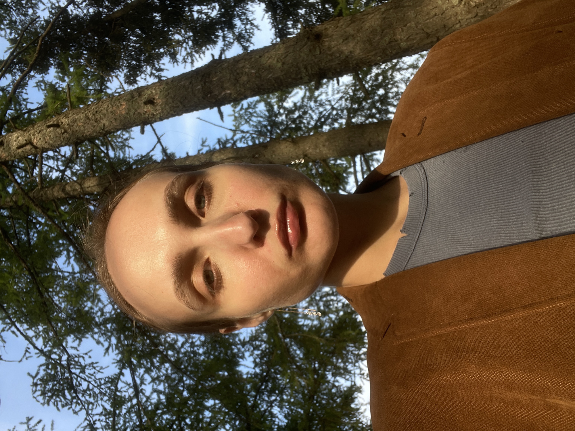 A young woman is photographed from a low angle, looking down at the camera, while positioned under trees. The background consists of tree branches and a glimpse of the sky. She is wearing a gray top with a brown jacket. Her face is illuminated by soft, natural light. The location is potentially Podshivalovo, Russia based on the city name provided. The image predominantly showcases the woman's face and the immediate natural environment surrounding her. No specific actions or interactions are discernible.