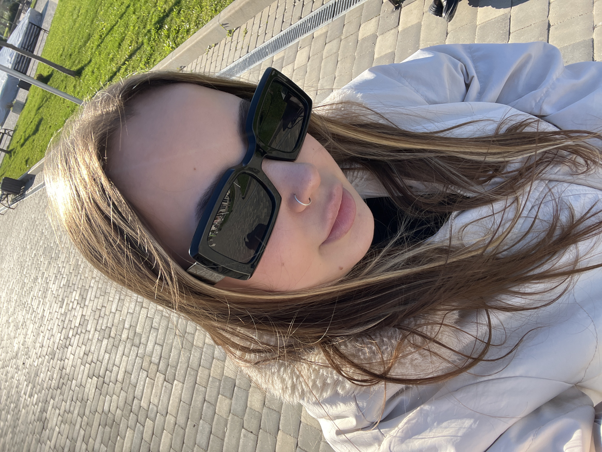 The image shows a person wearing black sunglasses and a nose ring. The person is wearing a white jacket, and has light brown hair with blonde highlights. The person is outside on a paved surface, likely a sidewalk or plaza. In the background, there is grass, benches, and some architectural structures. The image appears to be taken from a low angle, possibly a selfie. The location is Novopokrovskaya, Russia.