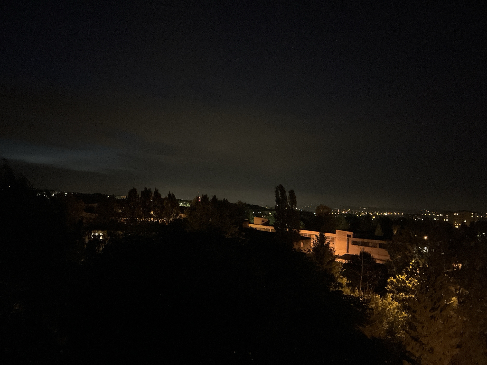 The image displays a nocturnal landscape captured from an elevated viewpoint. The foreground is dominated by a dark, silhouetted expanse of trees and foliage, appearing as deep shadows. In the mid-ground, a modern-style building, possibly light brown or beige, is illuminated by artificial light, showcasing its windows and architectural lines. Beyond this structure, a distant city or town stretches across the horizon, characterized by numerous scattered lights twinkling against the darkness, indicating urban development.
The sky is very dark, signifying nighttime, with faint, wispy cloud formations visible in the upper left and central areas, subtly catching light from below. A soft glow from light pollution emanates from the distant city, brightening the lower sky. No individuals or specific activities are visible. The scene portrays a serene, expansive view of a metropolitan area at night, observed from a quiet, natural vantage point. There is no visible text in the image.