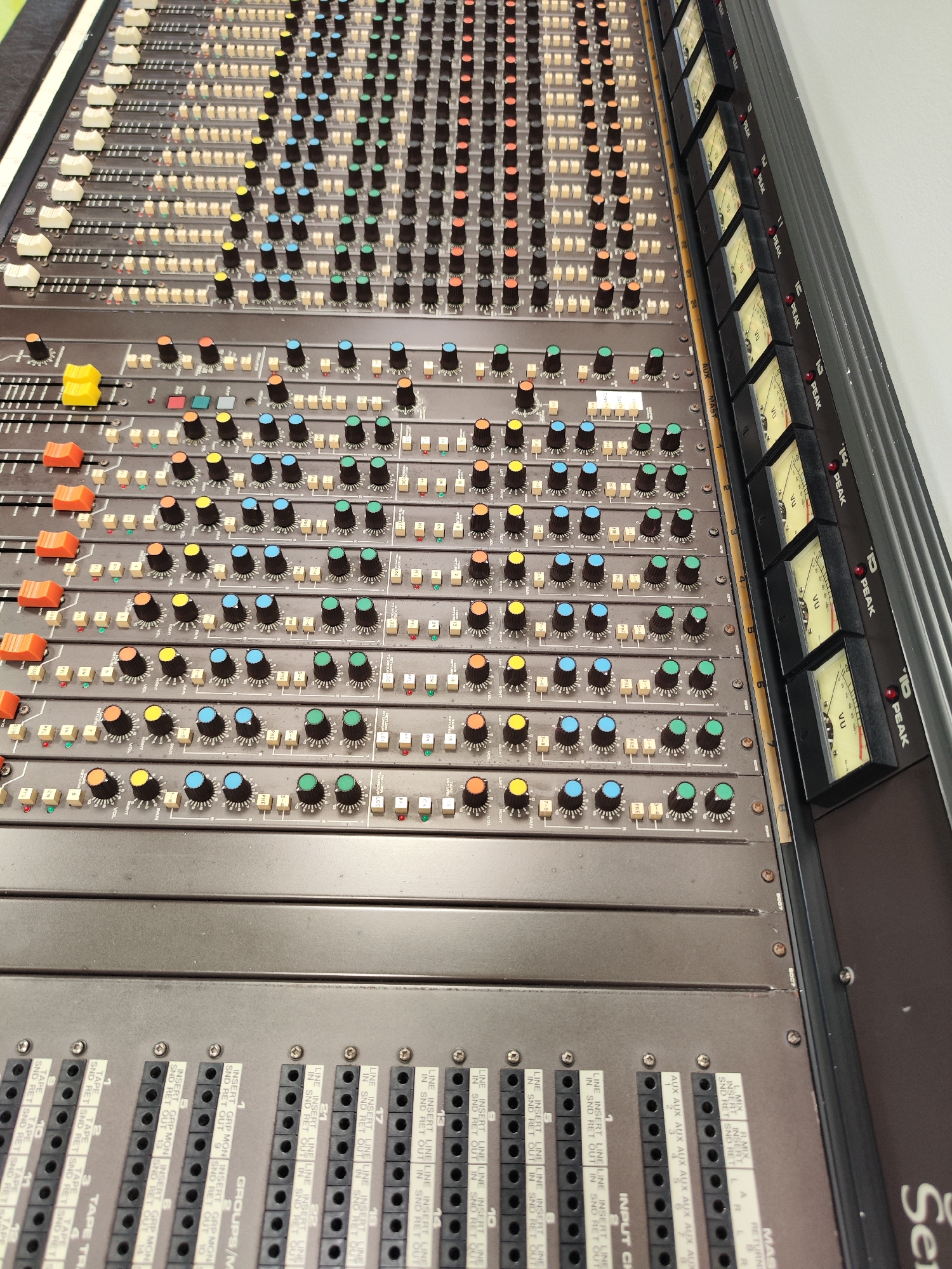 The image shows a close-up of a vintage audio mixing console. The console is brown/grey in color and features numerous knobs, faders, and switches of varying colors. VU meters are located on the right side of the console. Below the control surface are labeled input and output jacks for various channels, groups, and tape returns. The console's branding is partially visible in the lower-right corner.
There is no location information provided.