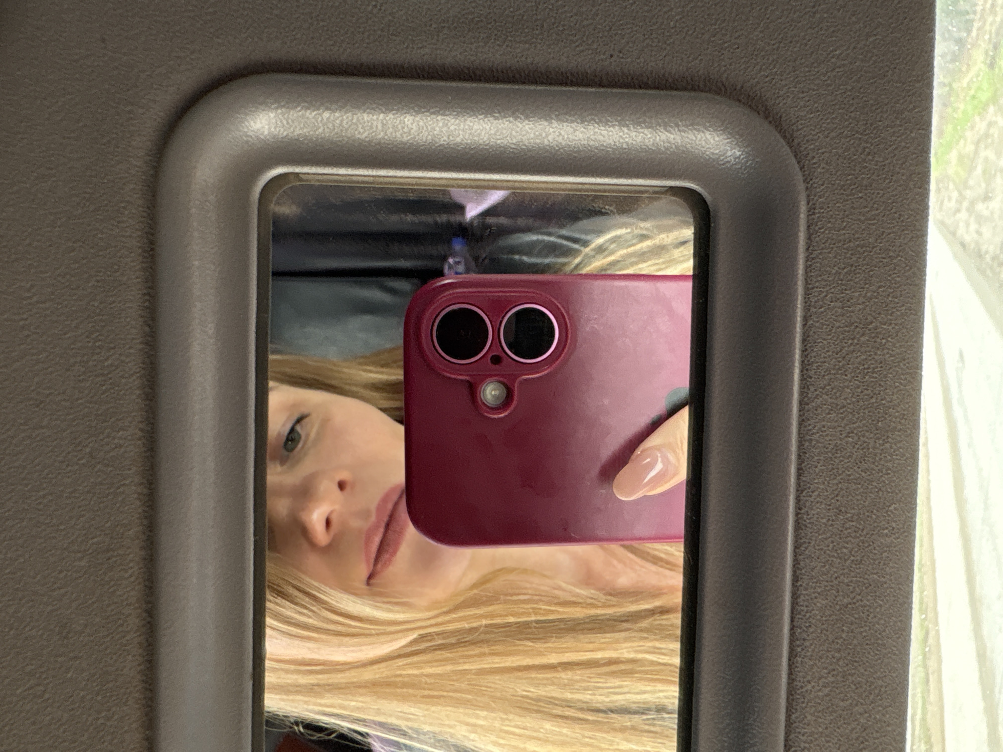 A close-up image shows a person taking a mirror selfie from inside what appears to be a vehicle. The central subject is a grey/silver framed mirror, reflecting the upper body of a person. The person has long, wavy blonde hair and a partially visible face, including one eye, nose, and lips with pinkish-brown lip makeup.
The person is holding a red or maroon smartphone, with its dual camera lenses prominently visible, pointed towards the mirror. A hand with light, manicured fingernails is seen holding the phone. Faintly reflected in the mirror above the phone is a small blue bottle, possibly a water bottle.
The setting appears to be the interior of a car, bus, or similar vehicle. The mirror is set into a grey, textured panel, likely a sun visor or part of the vehicle's ceiling or side panel. To the right of the mirror, a window frame is visible, showing blurred green foliage and bright light outside, suggesting daytime. No visible text is present in the image.
