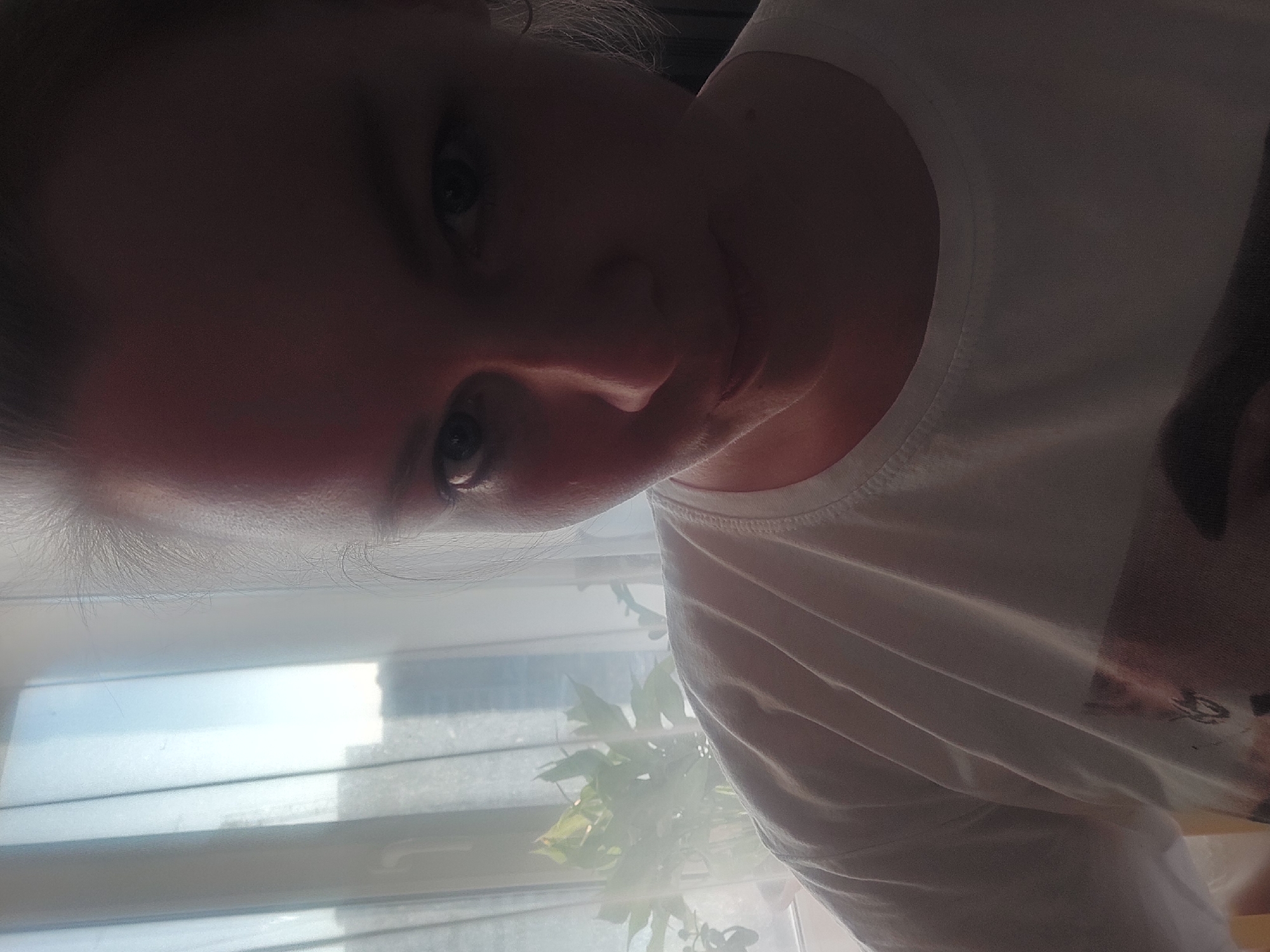 A medium close-up shot of a person indoors, possibly female, wearing a white t-shirt. The face is partially obscured by shadow. A window with blinds and a plant are visible in the background. The location may be Chaikovsky, Russia.