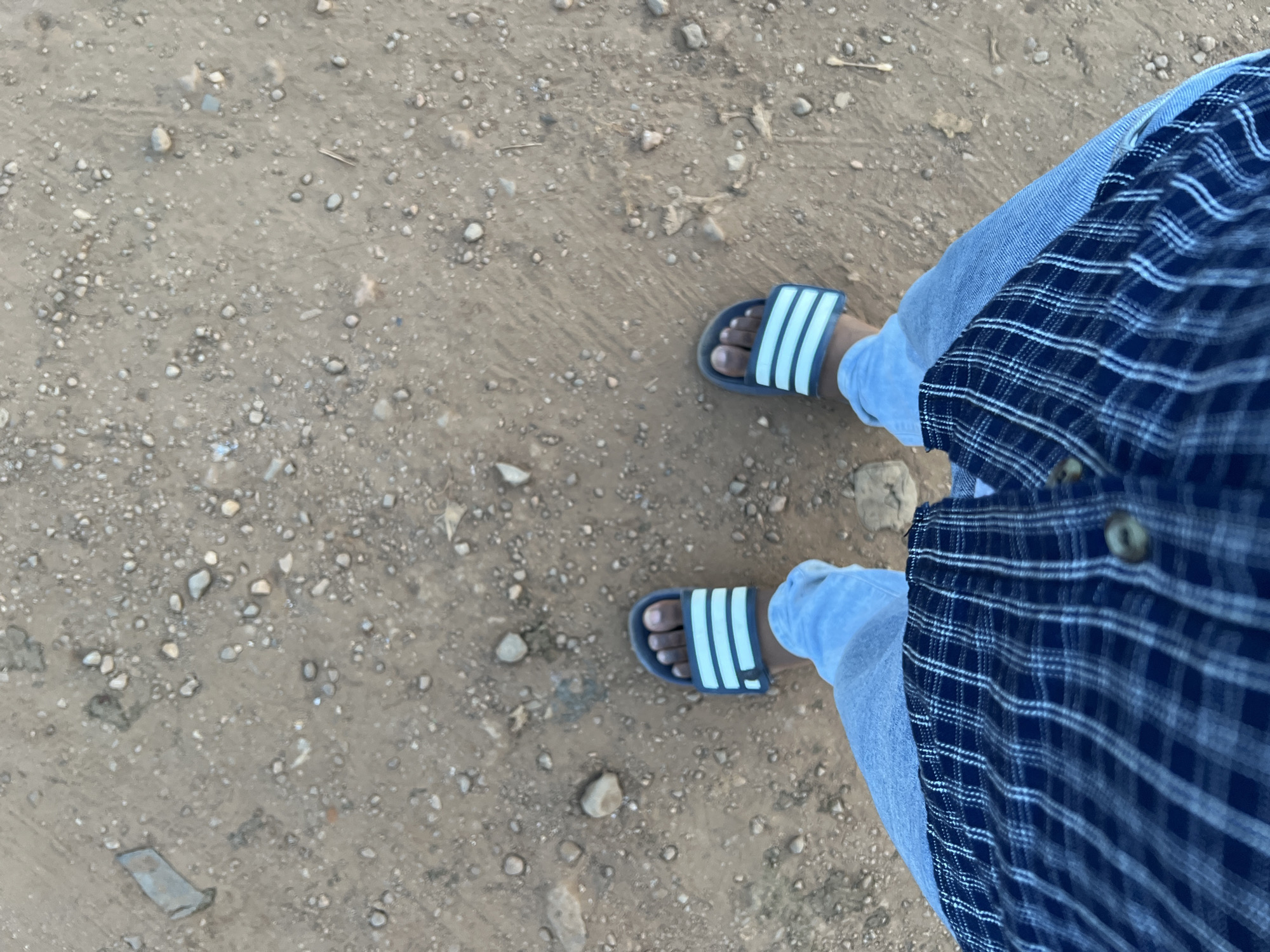 The image is a top-down view of a person standing on a dirt ground scattered with small stones. The person is wearing light blue jeans, a dark blue and white plaid shirt, and blue and white Adidas-branded slide sandals. The person is located outdoors on the ground, which may be a road. No specific location in Abuja, Nigeria, is discernible from the visual data provided.