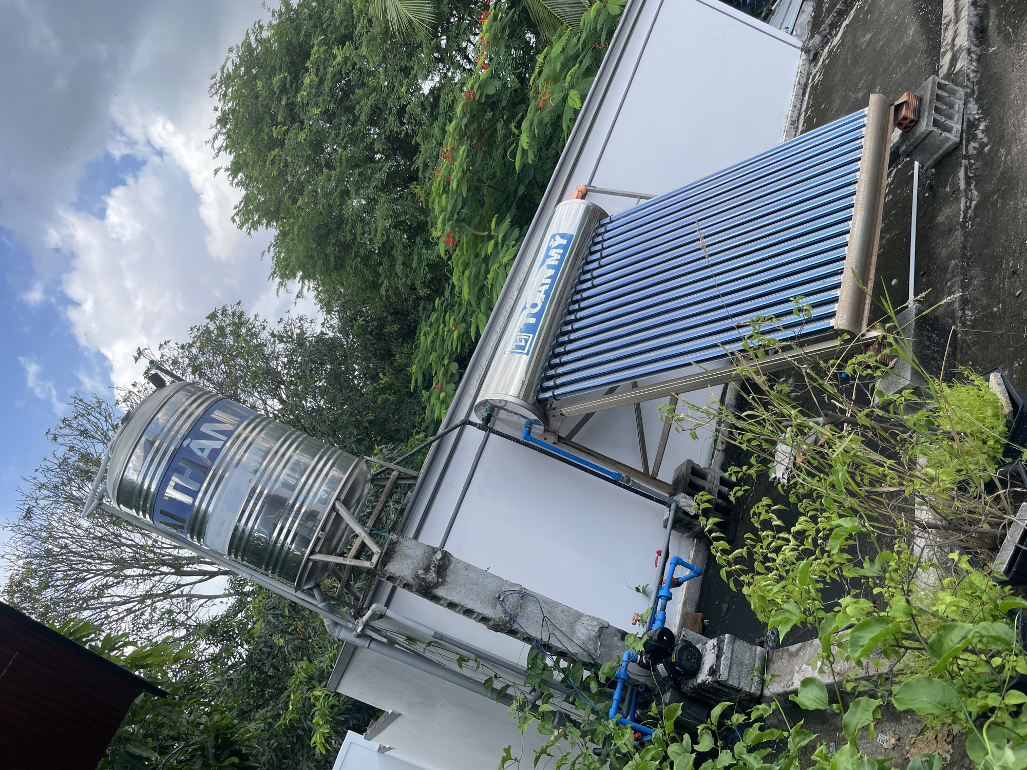The image depicts an outdoor scene on a rooftop, likely in Mũi Né, Vietnam, given the context. Dominating the view is a large, cylindrical stainless steel water tank mounted on a metal frame, angled upwards. Below it, a solar water heater with numerous blue vacuum tubes is installed on the rooftop. The rooftop itself appears to be made of concrete and shows signs of wear. Surrounding the equipment are various plants, some growing in planters and others encroaching on the structures, with lush green foliage. In the background, mature trees with dense green canopies are visible, and a glimpse of a white building wall is present to the left. The sky above is partly cloudy, with a mix of bright white clouds and darker, overcast areas. The overall impression is one of a functional, possibly residential, rooftop setup in a tropical environment.