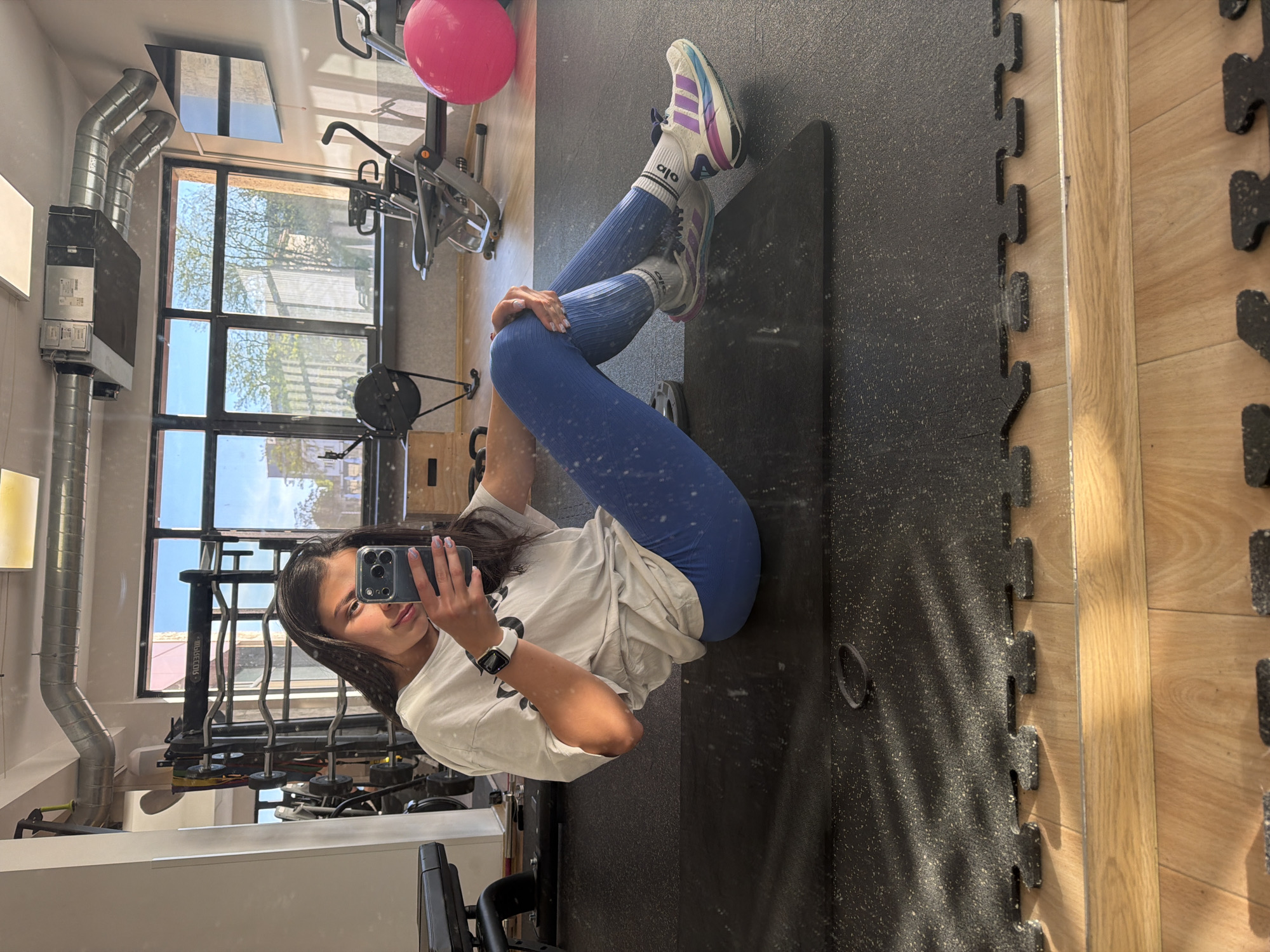 A young woman in a gym setting, possibly in Gabrovo, Bulgaria, given the location request. She is sitting on a black exercise mat, wearing blue leggings, a white oversized T-shirt, white socks with "alo" branding, and athletic shoes. She's holding a smartphone in her left hand, taking a selfie in a mirror. Her right hand rests on her knee. The background features gym equipment, a large window, and an industrial-style ceiling. The gym floor is made of interlocking black mats, with a section of light-colored wood flooring adjacent to the mats.