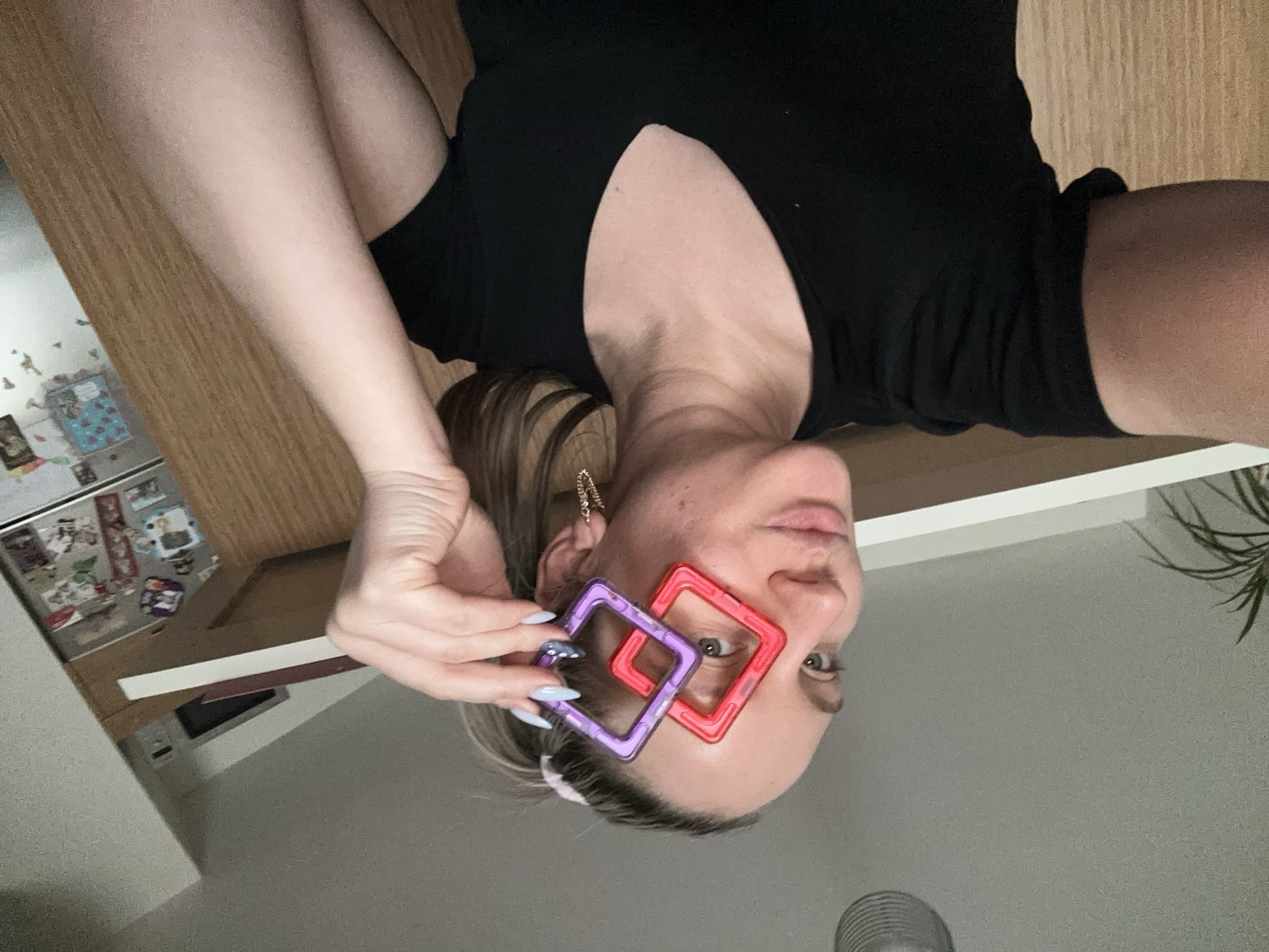 A woman is holding a purple and red square shaped object near her face. She has long, manicured nails. She has shoulder-length blond hair, and is wearing a black top. The background suggests she is in an apartment. The location is not discernible from the image. The city of Esenyurt is not discernible from the image.