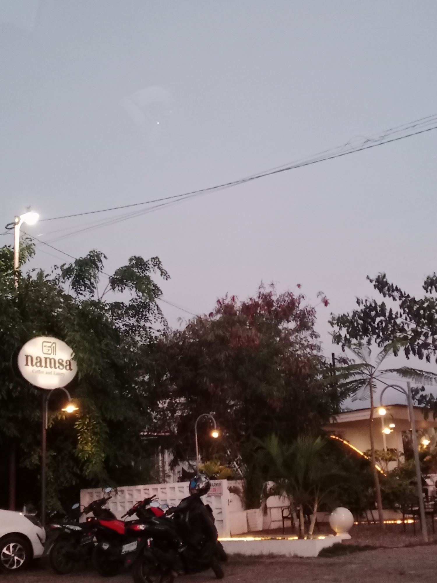 The scene is an outdoor area in Cotgapu, Indonesia at dusk. The sky is a pale grey, with an overhead power line visible. The "namsa" Coffee and Eatery sign is illuminated near the left of the frame. A white car is parked partially within the left side of the frame, along with several parked motorcycles. Behind the motorcycles, there's a white wall. Lush greenery, including trees and bushes, fills the midground, partly obscuring a building with lights on the right side. The environment appears to be a parking area near the cafe. A white spherical light is on the ground. A figure wearing a helmet stands next to one of the motorcycles.