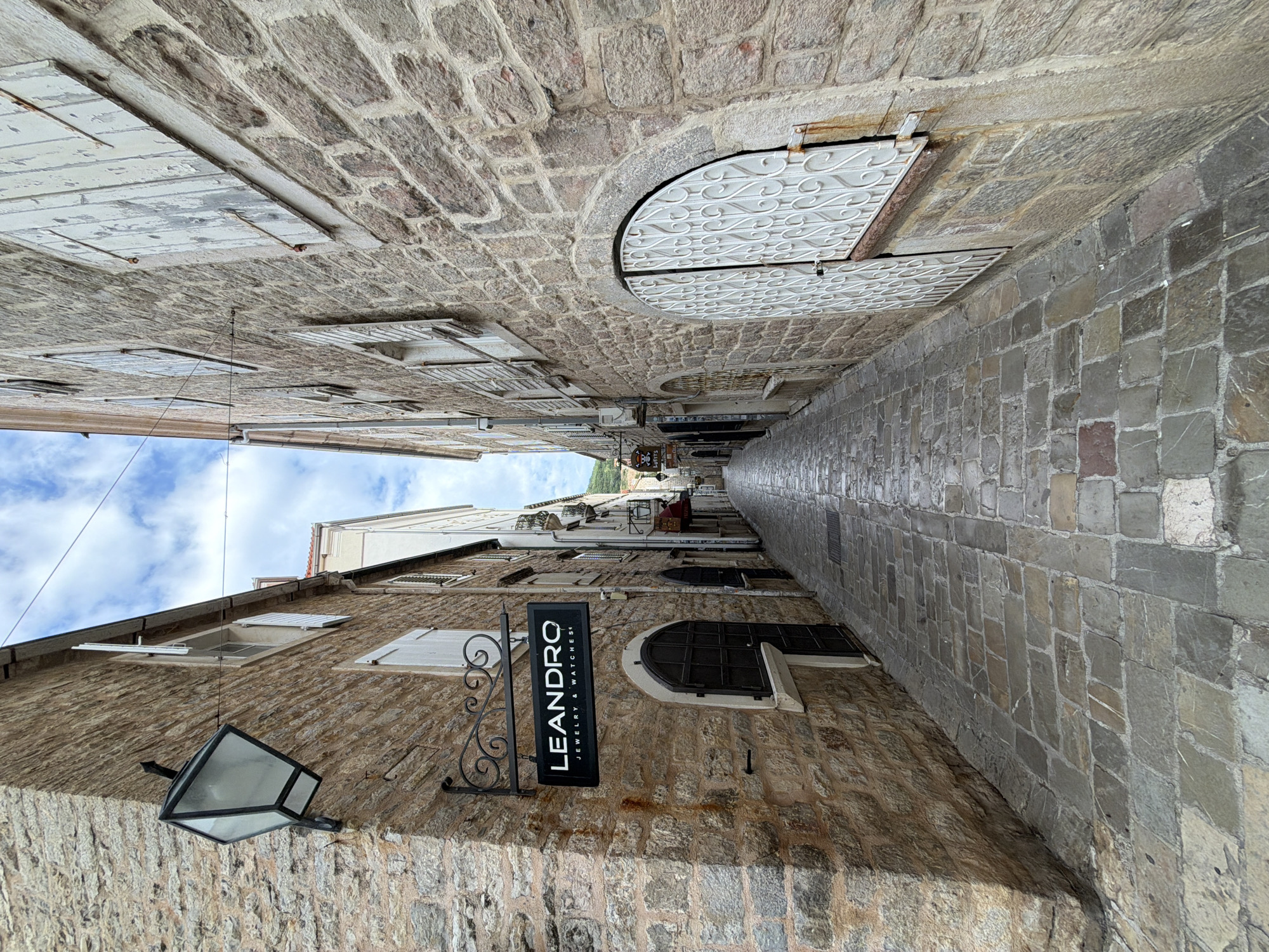 The image shows a narrow, stone-paved alleyway in Budva, Montenegro. The perspective is looking down the alley, with buildings on either side made of rough, tan stone. The sky, partially visible at the far end, is blue with scattered clouds. On the left building, there is a sign that reads "LEANDRO JEWELRY & WATCHES" and a hanging lantern. Above the sign, there are windows with white shutters, some open. The building on the right side has similar stone construction, with arched doorways and white metal covers over the windows. The pavement is made of irregularly shaped stone blocks, appearing smooth and worn. At the far end of the alley, there are hints of people and objects that suggest a commercial area.