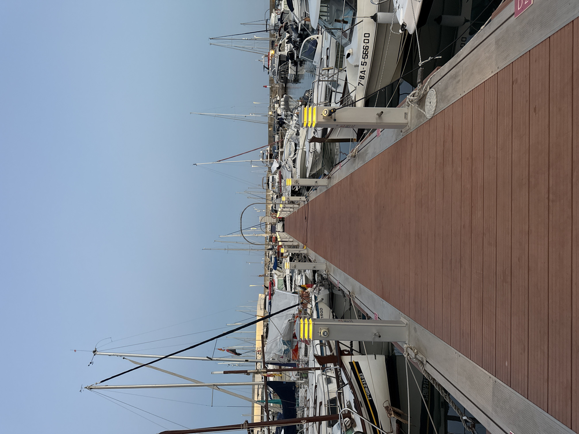 The image shows a perspective view of a wooden pier in a marina. The pier is lined with modern lampposts with yellow lights. On either side of the pier, numerous boats of various sizes are docked. The boats include sailboats with tall masts and motorboats. Many of the boats are white with some featuring dark accents or hulls. License plates are visible on some boats, such as "7° GIR 211-23" and "7° BA-5-566 00". The sky is a pale blue, suggesting either early morning or late evening. In the background, a larger structure, possibly a building or a bridge, with an arched element can be seen spanning across the marina. The water is calm. The location is identified as the Port Esportiu of Blanes, Spain. There are no people visible in the frame.
