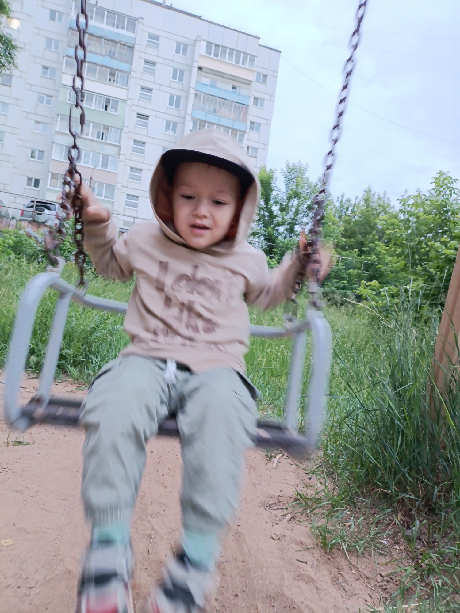 In the city of Chaikovsky, Russia, a young child is captured mid-swing on a swing set. The child, wearing a beige hooded sweatshirt and light green pants, is blurred due to the motion. They are holding onto the swing's chains, with their face turned towards the camera. The background features a tall, light-colored apartment building with rows of windows. Green trees and foliage surround the swing area, and the ground beneath is sandy with patches of grass. The sky is overcast.