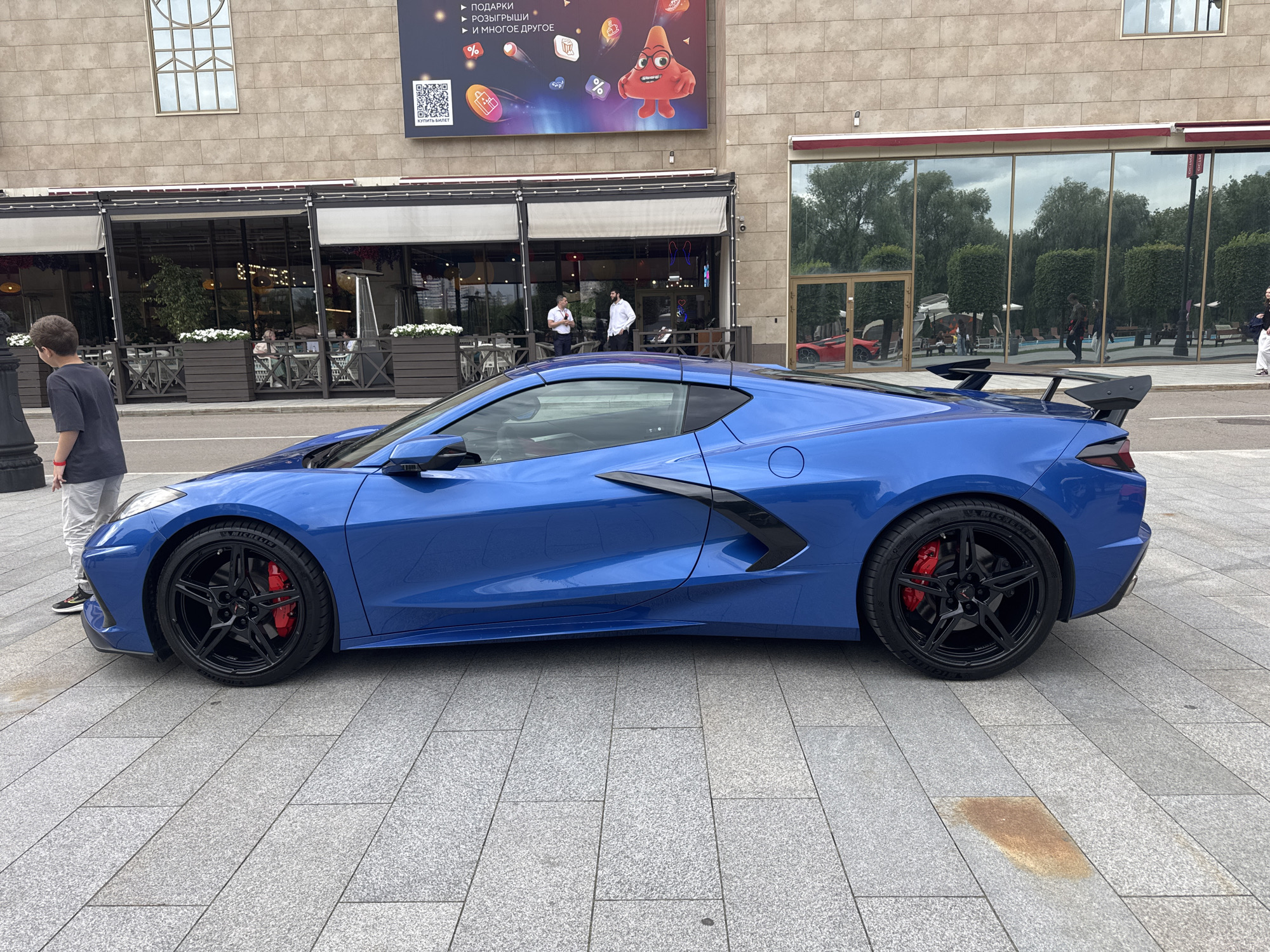 A bright blue Chevrolet Corvette C8 sports car is parked on a paved walkway in front of a modern building. The building features large glass windows reflecting trees and sky. A banner with graphics and Russian text is displayed above the entrance to the building, which appears to be a commercial establishment, possibly a restaurant or cafe, with outdoor seating visible through the windows. A young boy wearing a dark t-shirt and grey shorts stands to the left of the car, looking away from the camera. The car's rear spoiler is prominent. The walkway is constructed of large, light grey granite tiles. There is a red car visible through the building's reflection. The scene is outdoors, with overcast skies.