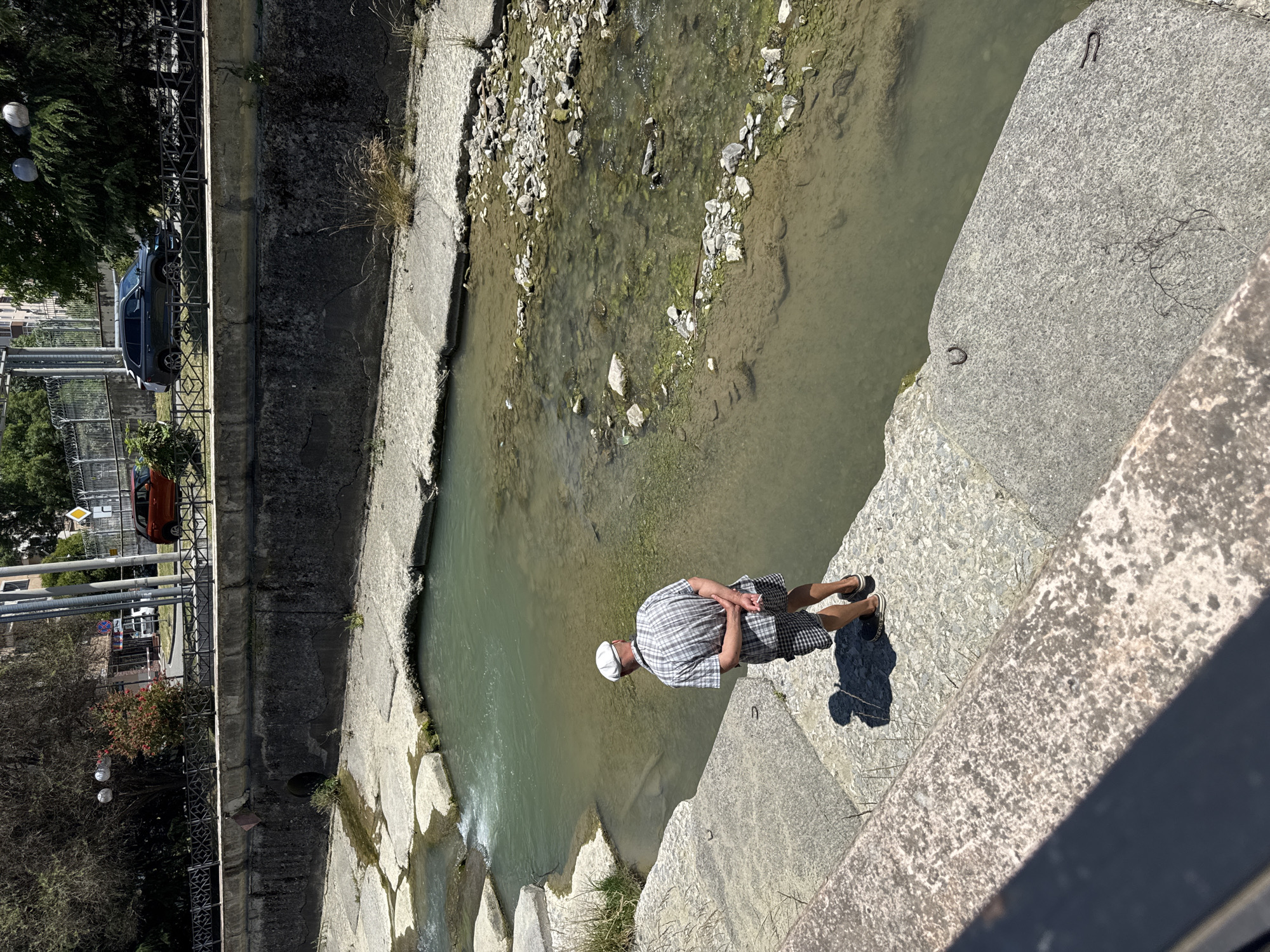 A man in a checkered shirt and shorts stands with his back to the camera, looking at a river in Sochi, Russia. He is wearing a white cap and holding a cigarette in his right hand. The river is shallow with a rocky bottom, and there are concrete structures along its banks. In the background, there are trees, buildings, and cars. The sun is shining, and it appears to be a warm day.