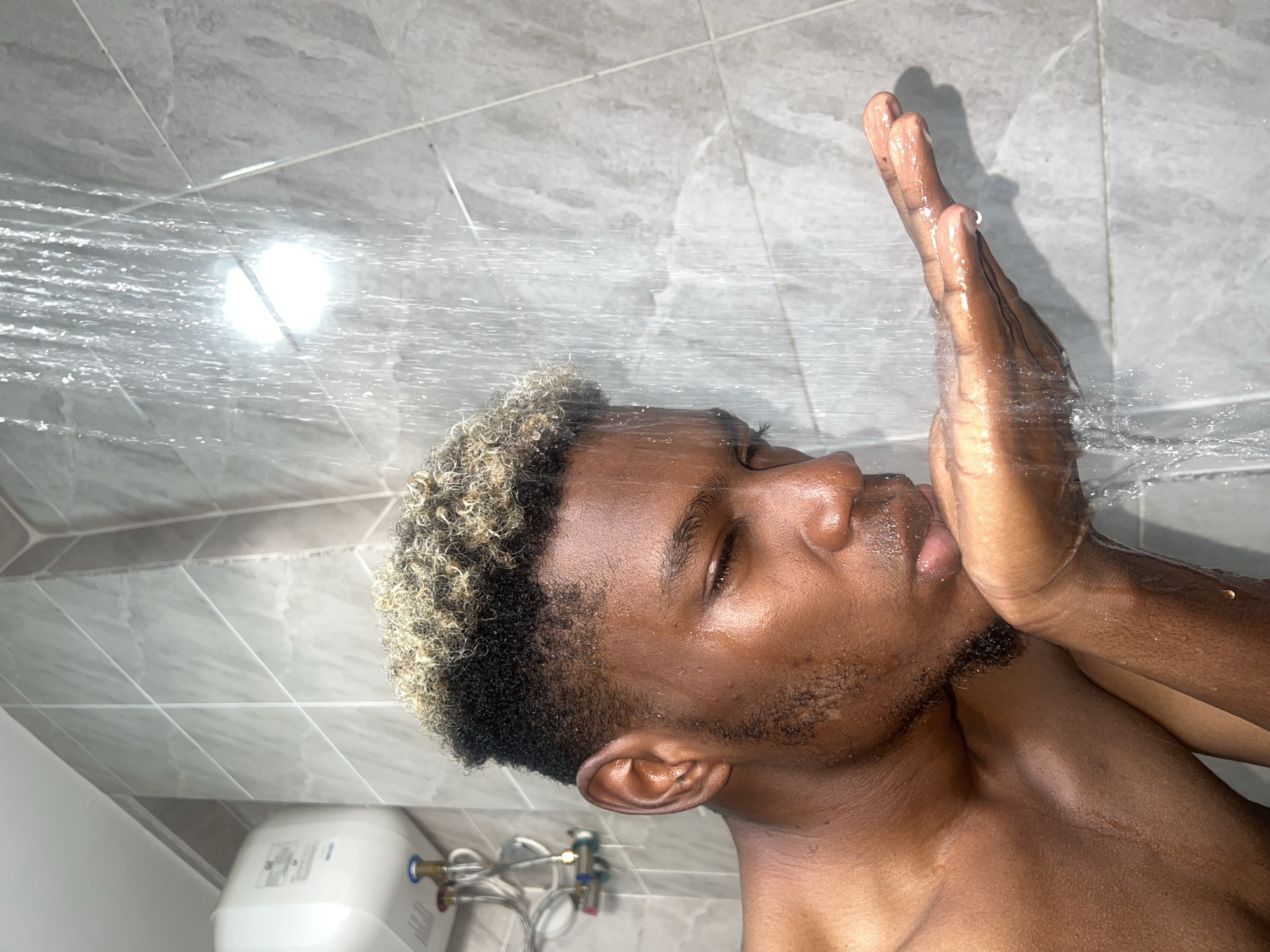 A medium close-up shot captures a young Black man showering in a bathroom, with the location provided as Igboefon, Nigeria. The image is rotated, showing the man diagonally across the frame, with his head towards the upper-left and his torso extending towards the lower-right. His eyes are closed, conveying a moment of concentration or relaxation. He raises his left hand to shield his face from the clear streams of water cascading from the upper-left of the frame and flowing diagonally downwards. Water droplets glisten on his wet dark skin and the light, possibly bleached, tips of his short, curly hair. He has short stubble or a faint beard along his jawline.
The background consists of light gray, rectangular tiled walls. In the lower-left corner of the image, a small white water heater or tank is visible. It is connected by exposed silver hoses and blue and red pipes, which are characteristic of plumbing fixtures. Another white bathroom fixture, possibly part of a toilet, is partially seen below the water heater. A bright light source creates a luminous reflection on the wet tiled wall and the water streams, indicating good lighting within the shower area. The text on the water heater's label is present but too obscured to be legible. The scene depicts a private act of personal hygiene.