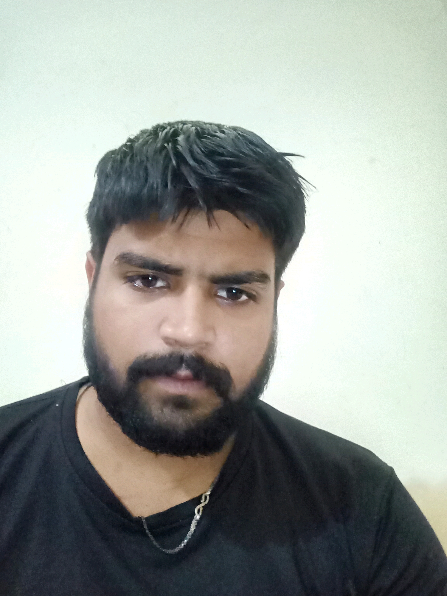 A medium shot portrait of a man with dark hair and a beard, wearing a black t-shirt and a silver necklace, is captured against a plain light-colored wall. The man's face is turned slightly towards the camera, with his eyes looking directly at the viewer. The lighting is soft, casting gentle shadows. The image appears to be a selfie, taken indoors. The scene is devoid of any specific identifying markers that would allow for a precise location within Dadri, India.