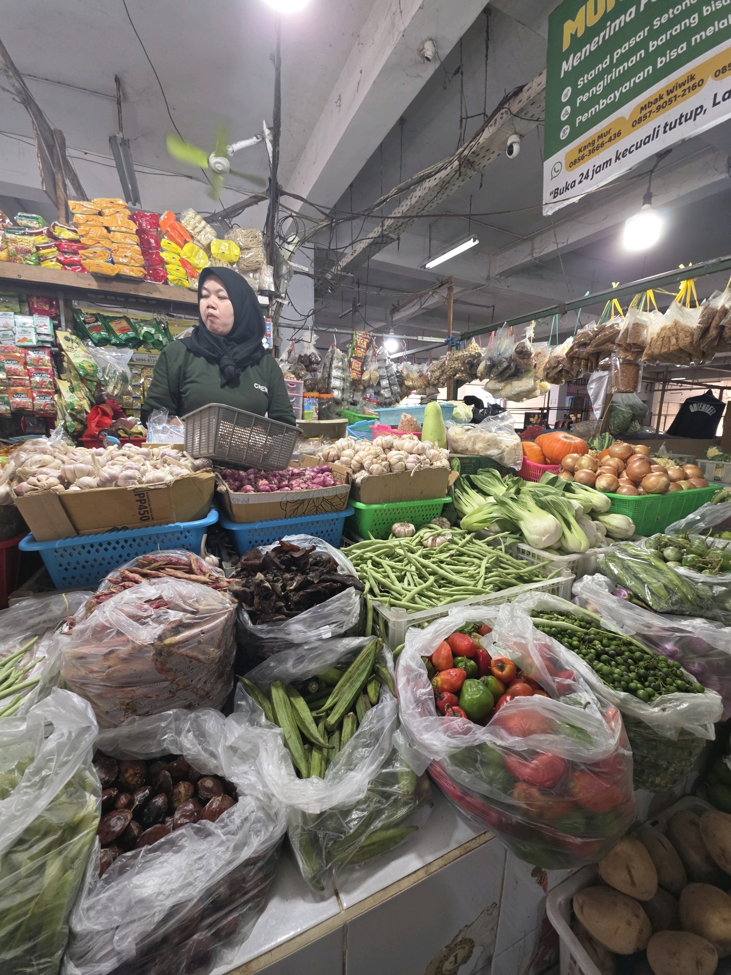 The image shows a market stall in the city of Unknown, Unknown. A woman wearing a black hijab and a green shirt stands behind a display of fresh produce. The stall is filled with various vegetables, including garlic, onions, green beans, bok choy, okra, tomatoes, bell peppers, and potatoes, arranged in baskets and plastic bags. Dried goods, such as snacks and possibly nuts or seeds, are hanging from the ceiling. A ceiling fan is visible, and overhead lighting illuminates the scene. A surveillance camera is mounted on a metal beam above the stall. A green sign with white text, written in Indonesian, is also visible, providing information about the market's services and contact details. The overall atmosphere suggests a bustling marketplace.