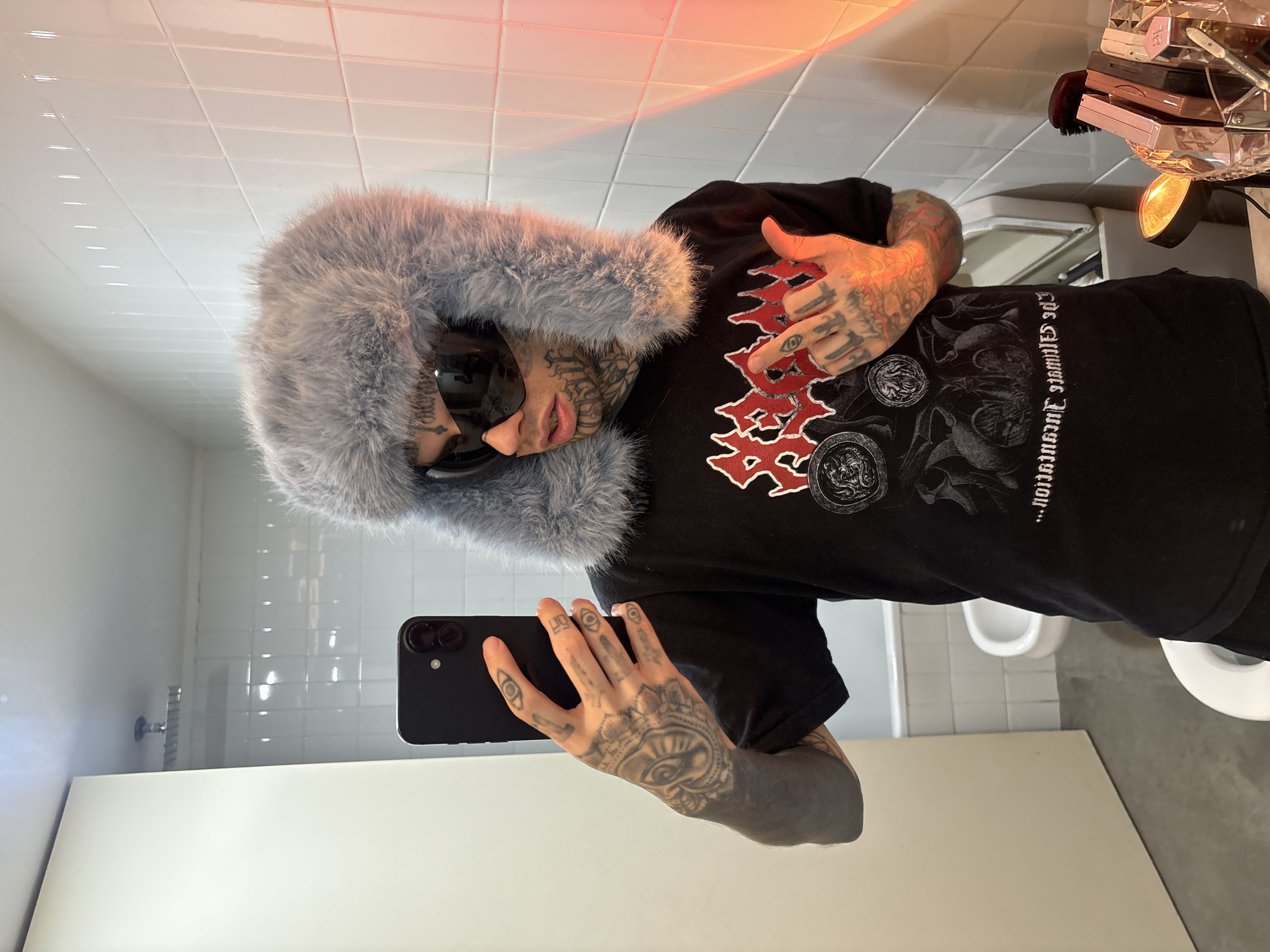A tattooed person wearing a fur-lined hat and sunglasses takes a selfie in a bathroom. The person is wearing a black t-shirt with a graphic design. The bathroom has white tiled walls, a toilet, a sink, and a countertop with various items, including makeup. The location is possibly in Buenos Aires, Argentina.