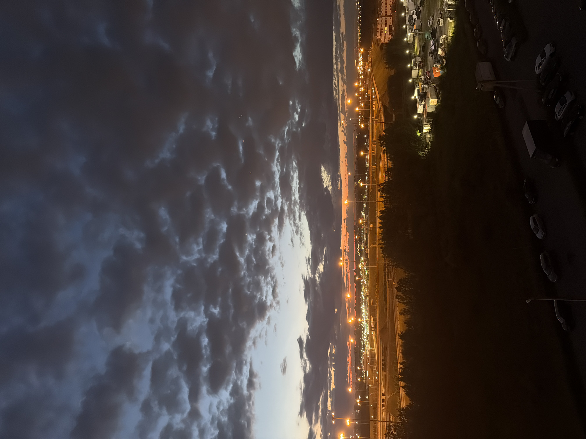 The image depicts an aerial view of a city at dusk, with a dramatic sky filled with dark, textured clouds. The sky transitions from a deep twilight blue at the top to a warm orange and yellow hue near the horizon, illuminated by the setting sun. Below the sky, a complex network of elevated highways and roads is visible, crisscrossed by streetlights that cast a warm glow on the asphalt. A dense line of trees forms a dark silhouette between the highway and the foreground. In the middle ground, a parking lot is filled with various vehicles, including trucks, trailers, and cars, with some lights visible from nearby buildings. The foreground is darker, with more parked cars and a single streetlight. The scene conveys a sense of urban sprawl and the transition from day to night, with artificial lights beginning to dominate the landscape.