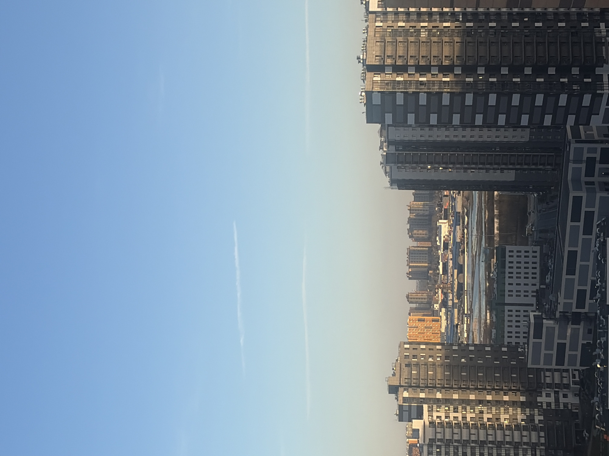 The image presents an urban landscape, likely Saint Petersburg, Russia, under a clear, bright blue sky. The view is from a high vantage point, looking across a sprawling city.
The upper portion of the frame features an expansive, unblemished blue sky, transitioning to a lighter, somewhat hazy tone towards the horizon line where it meets the cityscape. Two faint, white contrails are visible, stretching diagonally across the sky.
The lower part of the image is filled with a dense array of modern, multi-story buildings. These structures primarily exhibit dark or gray facades, with repeating patterns of windows, balconies, and architectural elements. The buildings appear to be residential or commercial, varying in height and design, contributing to a contemporary urban aesthetic.
In the mid-ground, a body of water, possibly a river or canal, is visible, winding through the urban expanse. Its surface reflects the sky, and along its banks, further low-rise buildings and industrial-looking areas can be observed. A distinct cluster of buildings in the distance glows with a warm, golden-orange hue, contrasting with the predominantly cooler tones of the other structures.
The lighting suggests a time of day with the sun relatively low, possibly late morning or late afternoon, as some buildings are brightly illuminated while others cast subtle shadows. The distant cityscape appears slightly hazy, characteristic of an urban atmosphere. There are no visible people or discernible text in the photograph.