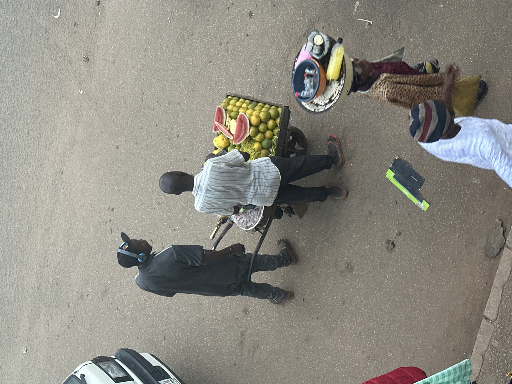 In the city of Bukuru, Nigeria, on a paved road, two individuals are seen in the foreground. The person on the left, wearing a dark grey t-shirt, patterned trousers, and black sandals, has blue headphones on and is looking towards the right. To their right, a person wearing a striped shirt and black trousers stands by a cart laden with oranges and slices of watermelon. Further to the right, a woman walks carrying a tray of items, including plastic bottles, on her head, dressed in a leopard-print jacket. Another person in a white garment and a striped hat walks in front of her. A portion of a white vehicle is visible on the left edge of the frame. The environment is an outdoor, urban setting with the texture of asphalt and some scattered debris on the ground.