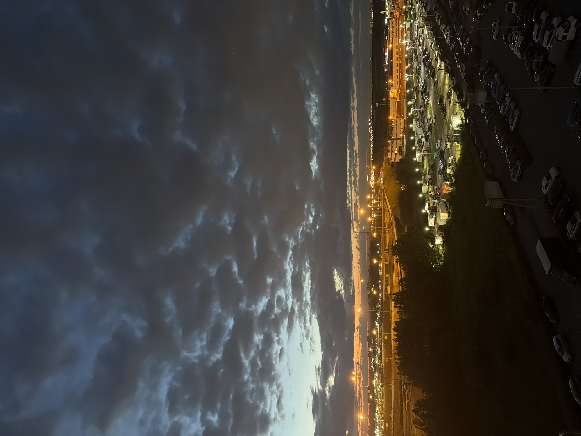 The image depicts an aerial view of a parking lot at dusk. The sky is filled with dark, heavy clouds, with hints of orange and yellow peeking through at the horizon, suggesting the last light of sunset. Below the sky, numerous streetlights illuminate a vast, densely packed parking lot filled with many cars, mostly dark in color. Some trucks and other vehicles are also visible within the parking area. In the mid-ground, a dark green treeline separates the parking lot from a complex of low-rise buildings, also lit by artificial lights. A network of roads, possibly a highway or freeway, with more streetlights, is visible in the distance, connecting to the illuminated area. The overall scene conveys an urban or industrial landscape at the end of the day.